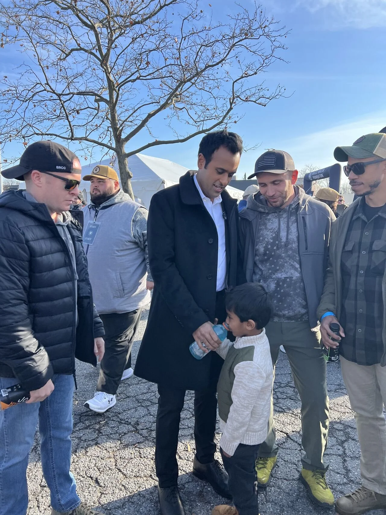 A group of men and a boy gathered outdoors under a leafless tree on a clear day. The men are wearing casual and outdoor clothing, and one man is holding a water bottle for the boy, who is drinking from it. They seem to be engaged in a conversation.