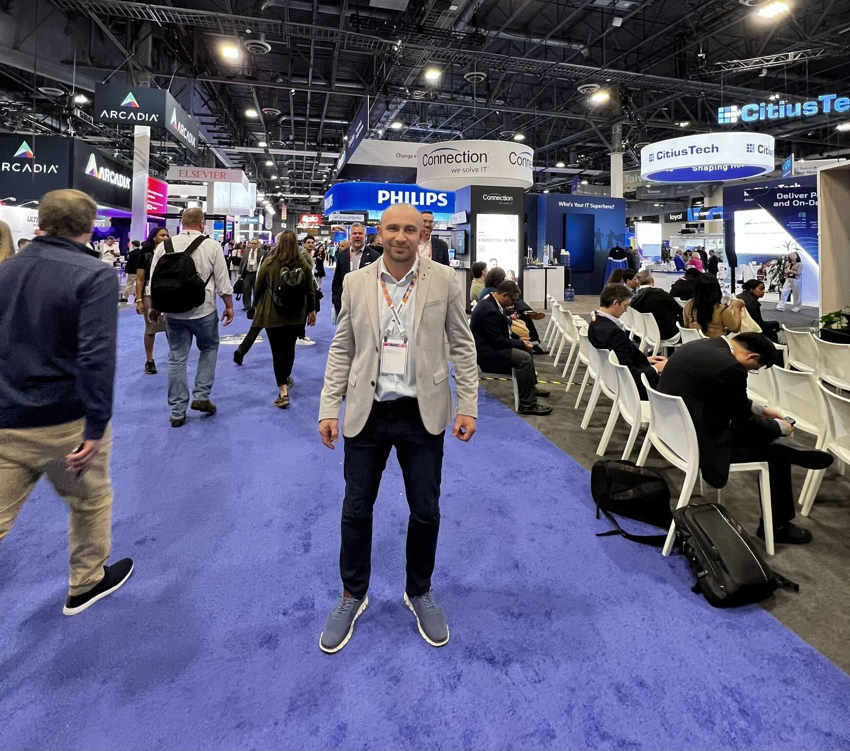 Man standing on blue carpet at a busy technology conference, surrounded by attendees and numerous exhibition booths with company logos and displays.