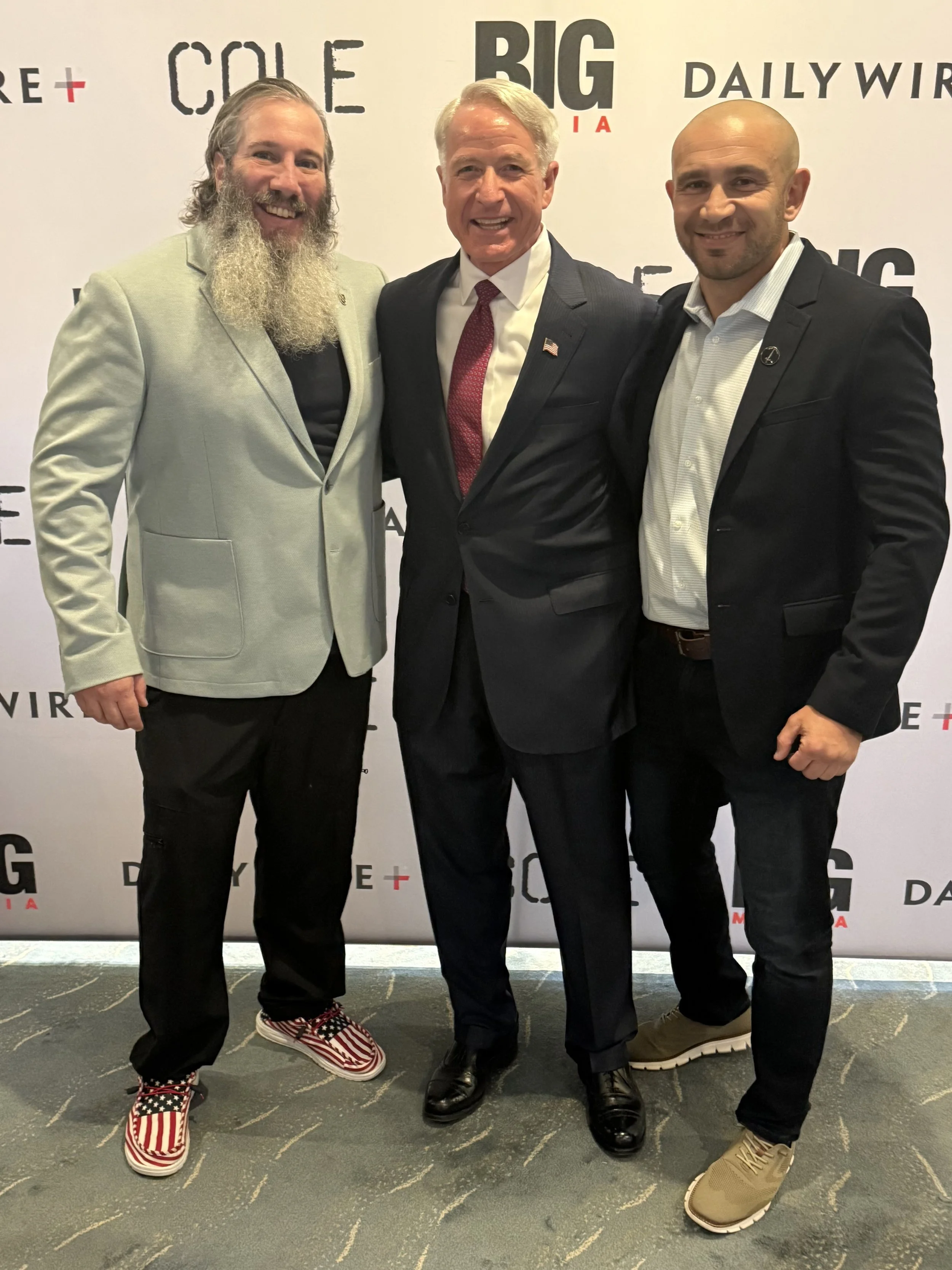Three men in suits smiling and standing together in front of a backdrop with the logos of Comedy Central, Big USA, Daily Wire, and more.