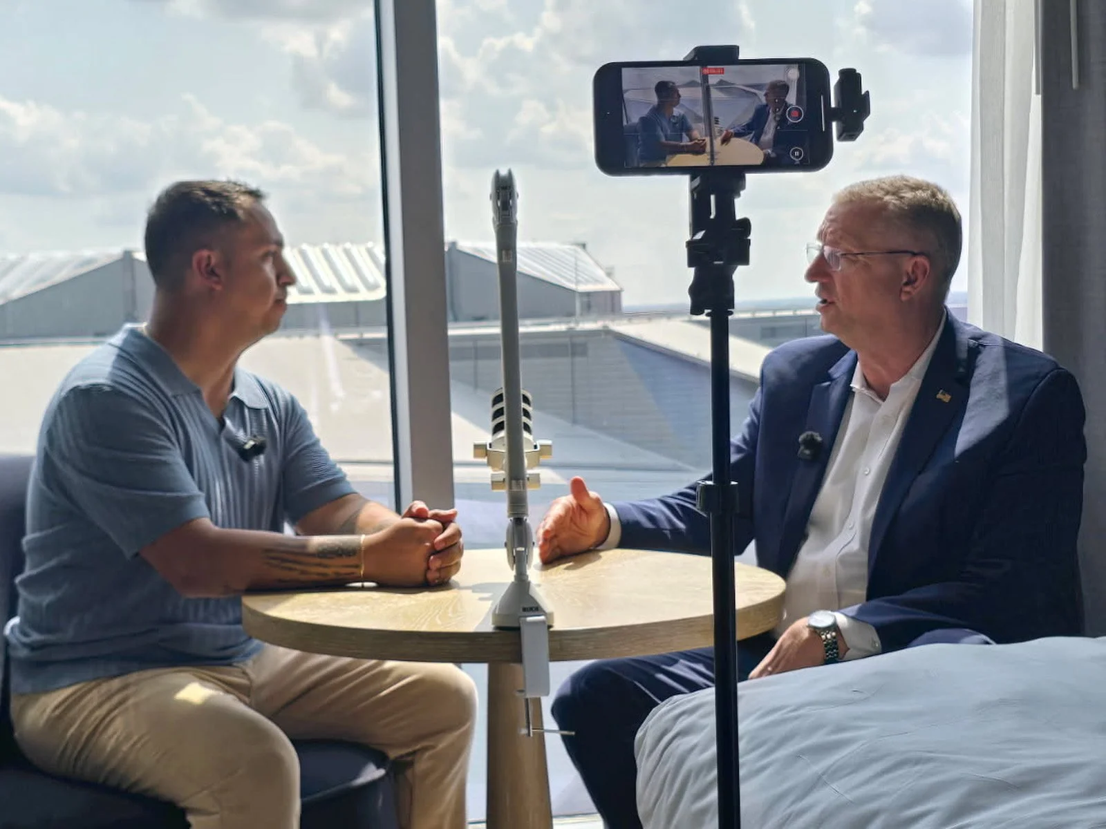 Two men sitting at a round table, holding hands, being recorded on a smartphone mounted on a tripod, with a large window showing the sky in the background.