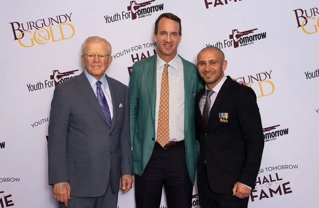 Three men standing together at an event with a step and repeat banner in the background. The banner features logos and text for 'Burgundy & Gold,' 'Youth for Tomorrow,' and 'Hall of Fame.'