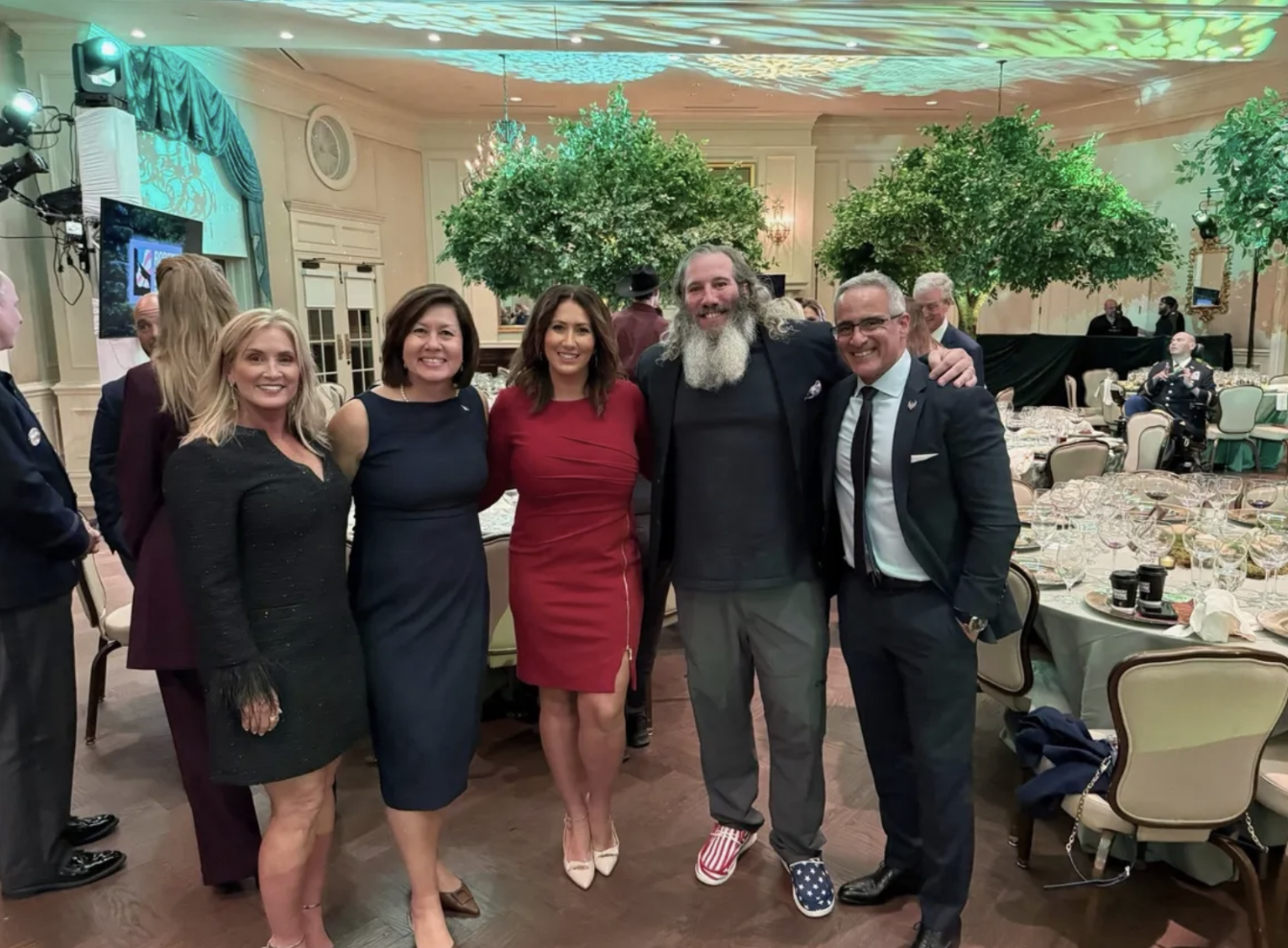 Group of five people, three women and two men, posing together at a formal event in a decorated banquet hall with trees and elegant table settings.