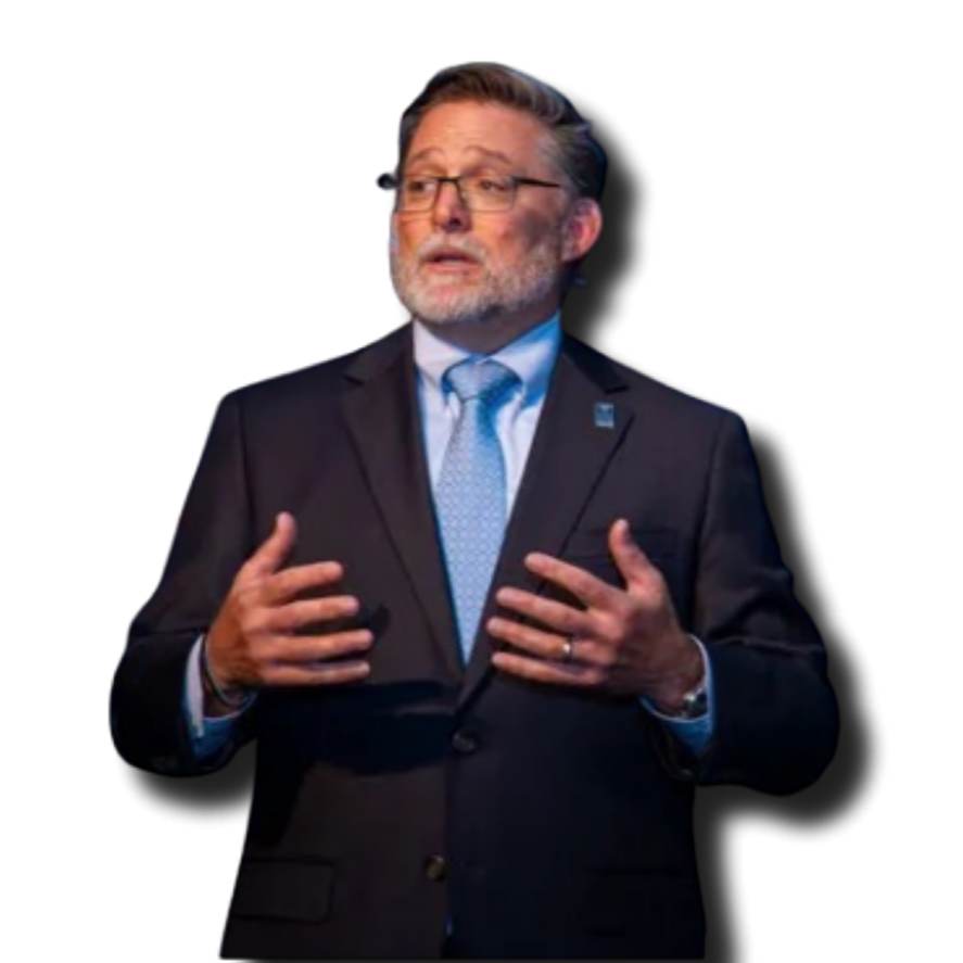 A middle-aged man with glasses, a beard, and gray hair speaking or presenting, dressed in a black suit, white shirt, and light blue tie, against a black background.