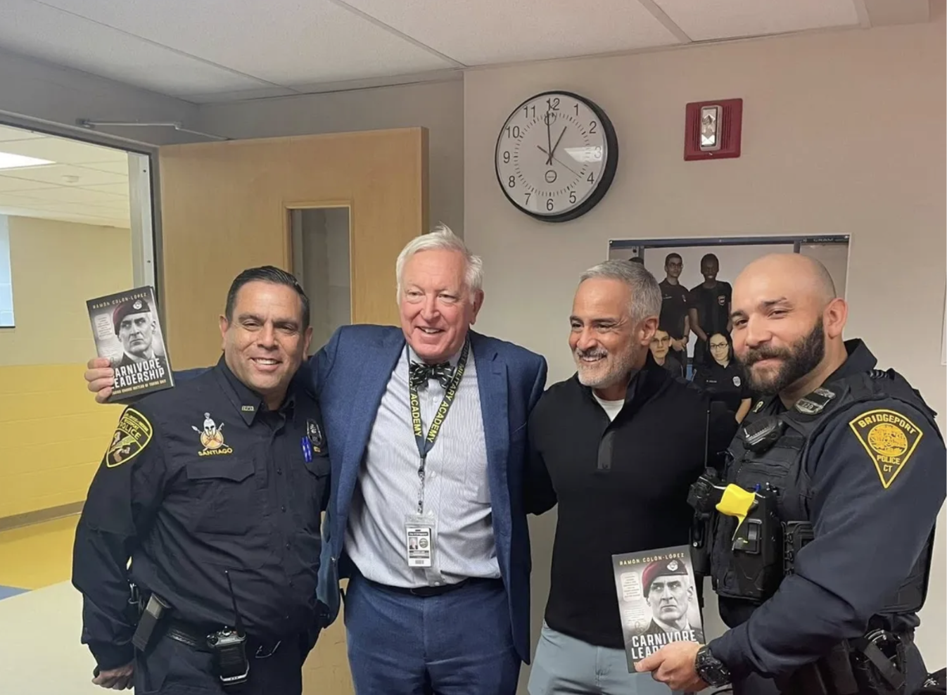 Four men, including two police officers, standing inside a room with yellow walls, smiling and posing for a photo. Two of them are holding copies of a book titled 'Carnivore Leadership,' featuring a man wearing a red beret on the cover. There is a cl