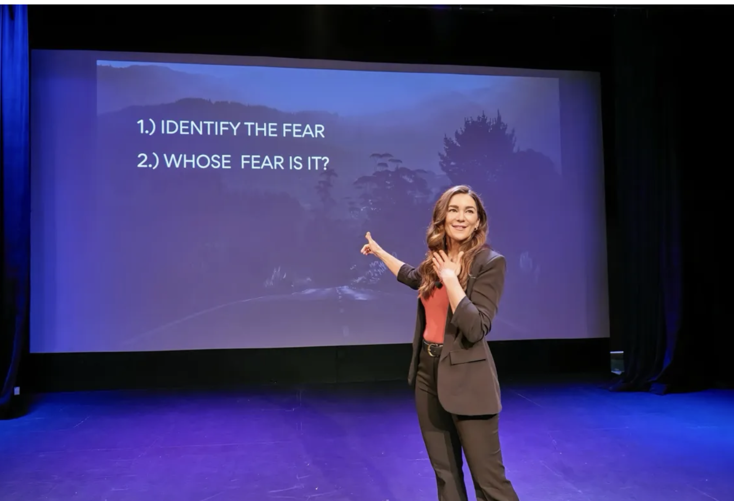 A woman in professional attire standing on a stage with a large screen behind her. The screen displays the text '1.) IDENTIFY THE FEAR 2.) WHOSE FEAR IS IT?' in white font against a background of trees and mountains.