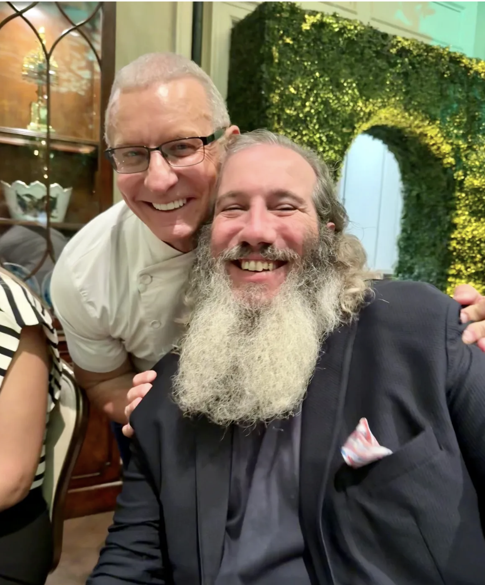 Two smiling men, one with glasses and a white chef's coat, and the other with a long beard and dark suit, hugging each other at a social event with green floral decor in the background.