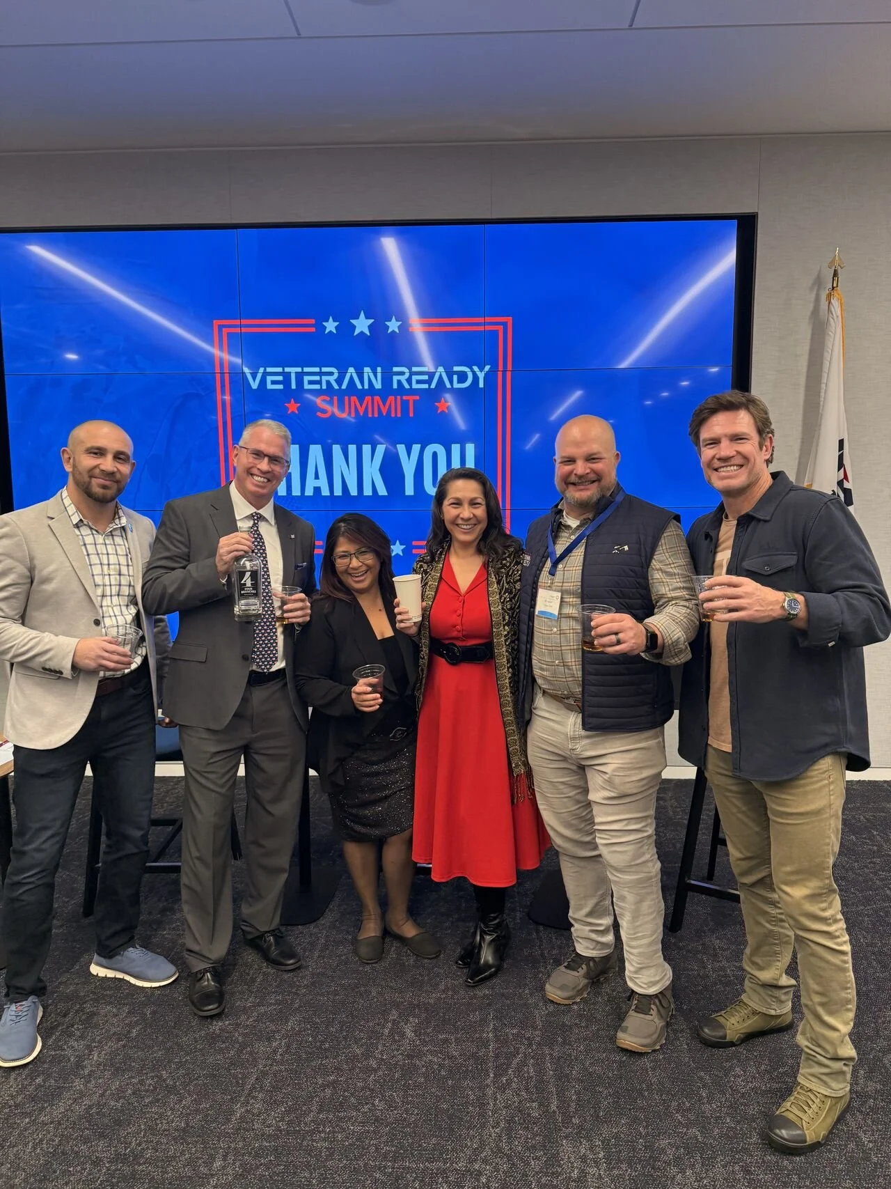 Group of six people standing together, smiling, holding drinks, at an event titled 'Veteran Ready Summit' with a large digital screen behind them displaying the event's name and a thank you message.