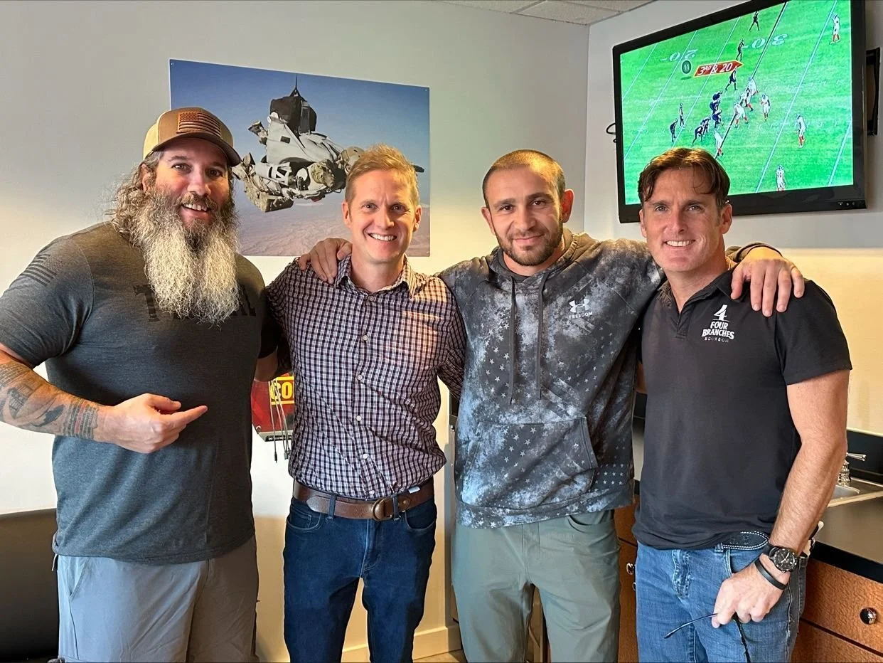 Four men standing together indoors, smiling, with one pointing at his arm. Behind them is a wall art of a vehicle and a television screen displaying a football game.