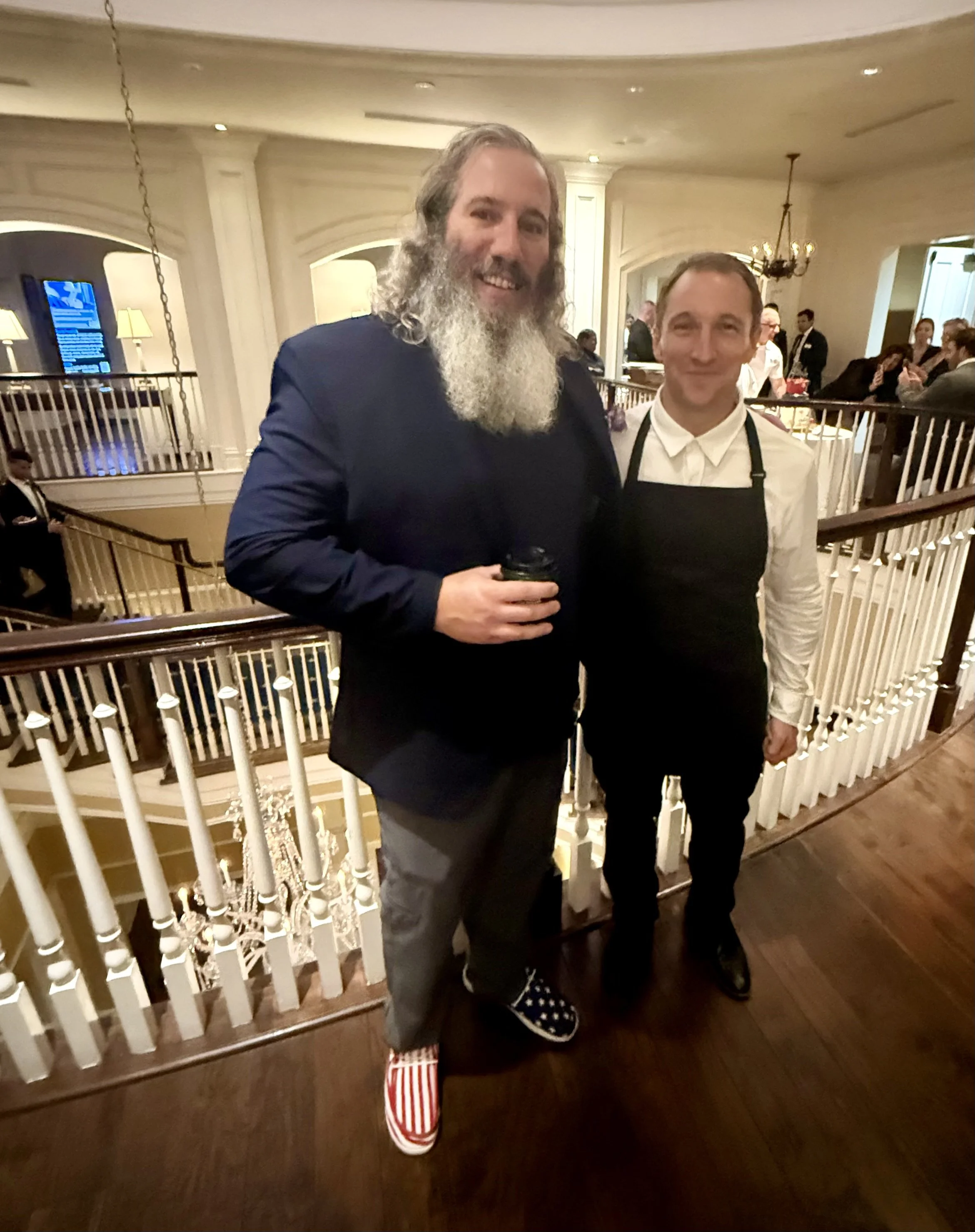 Two men smiling and posing for a photo in an elegant, well-lit room with a chandelier, white railings, and people in the background. One man has a long beard and is holding a drink. The other man is wearing a white shirt, black apron, and polka-dot s