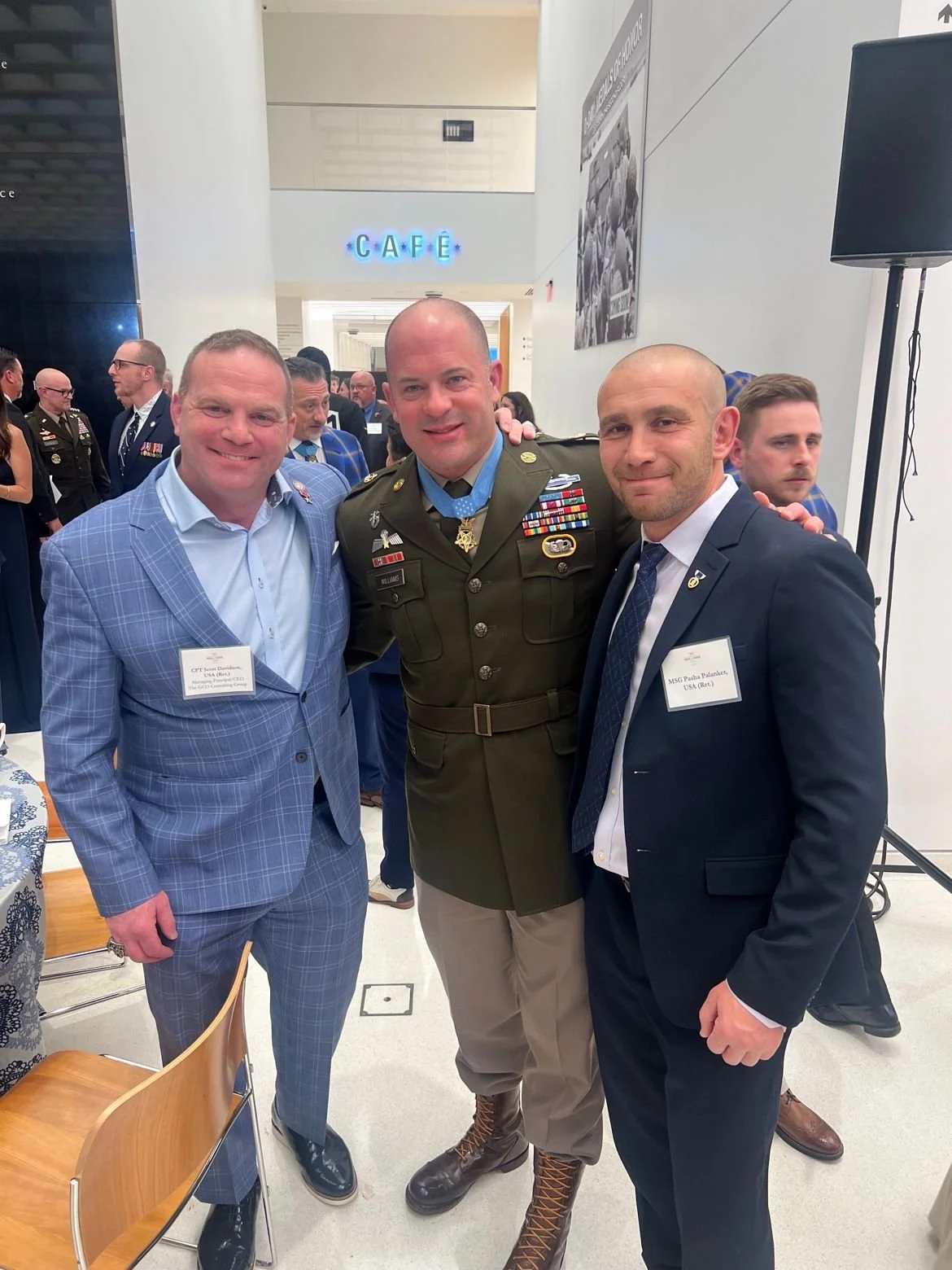 Three men dressed in suits and military uniform standing together at an indoor event, smiling for the photo, with a group of people and a cafe sign in the background.