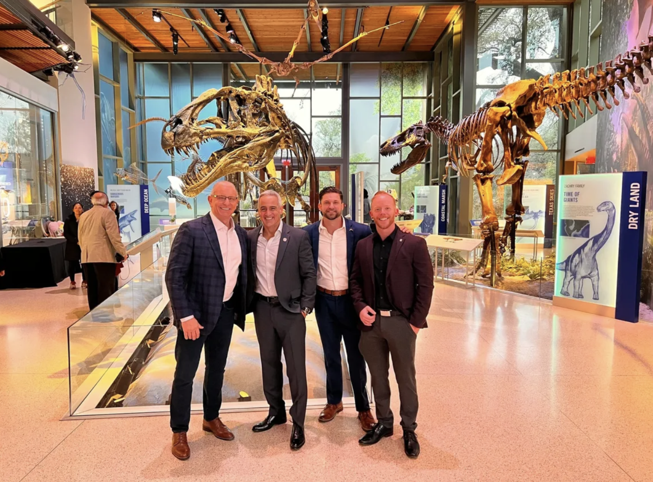 Four men in suits and business attire stand together smiling in front of dinosaur skeletons inside a museum exhibit, with large windows and informational displays about prehistoric worlds.