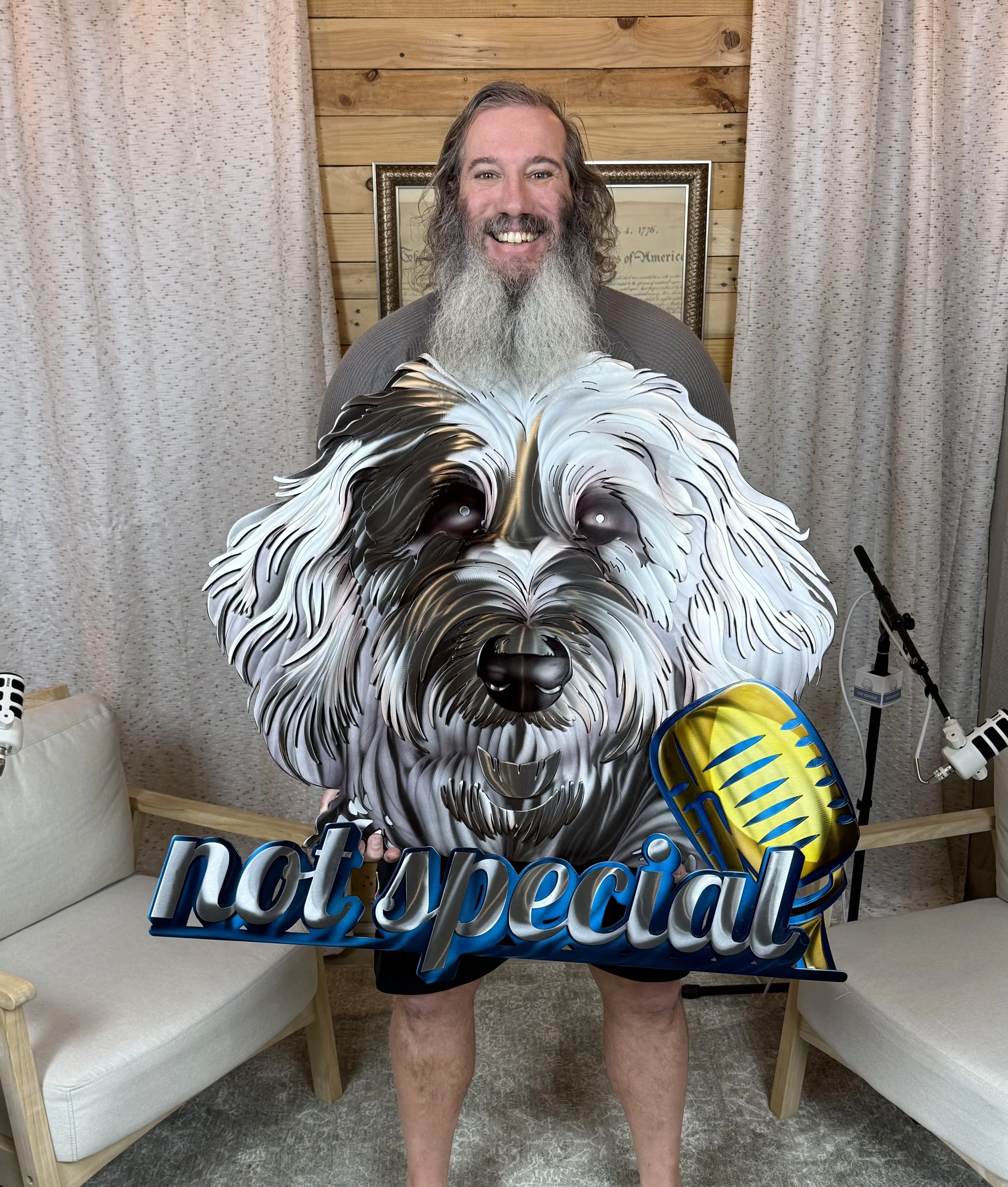 A man with a long gray beard and mustache holding a large dog-shaped sign that says "protected" in blue 3D letters, standing in a room with a wooden wall backdrop and beige curtains, with a beige sofa and a chair in the background.