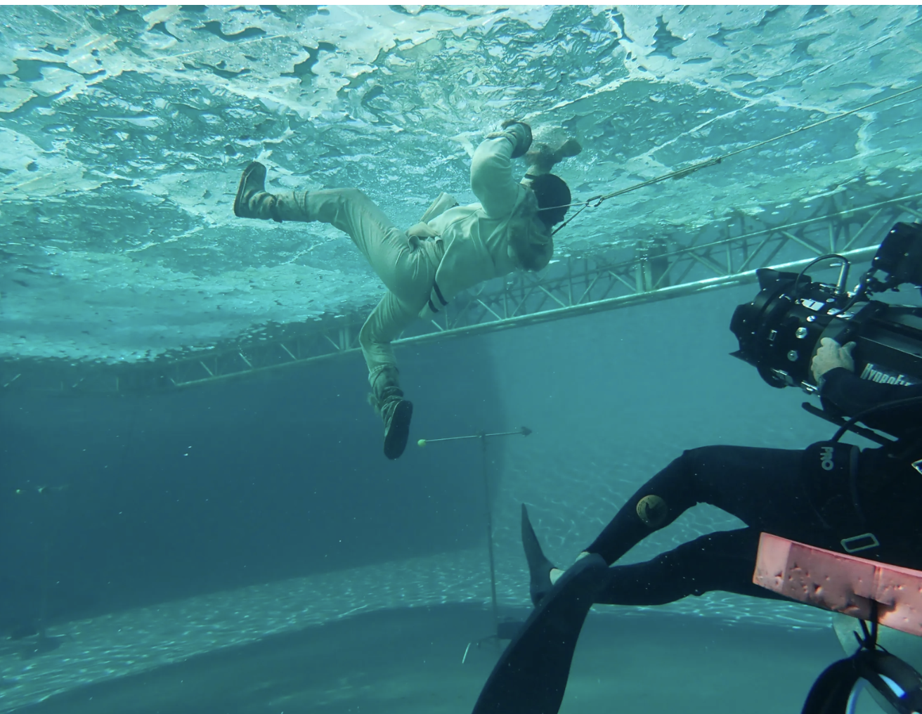Underwater view of a person in a suit, pulled by a cable, with a diver operating a camera nearby.