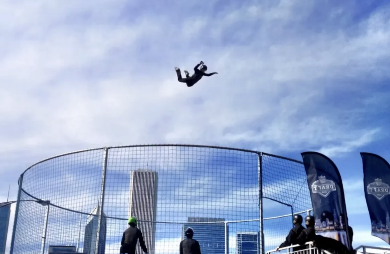 A person is mid-air doing a stunt above a trapeze net, with several people below watching, against a background of tall buildings and a partly cloudy sky.