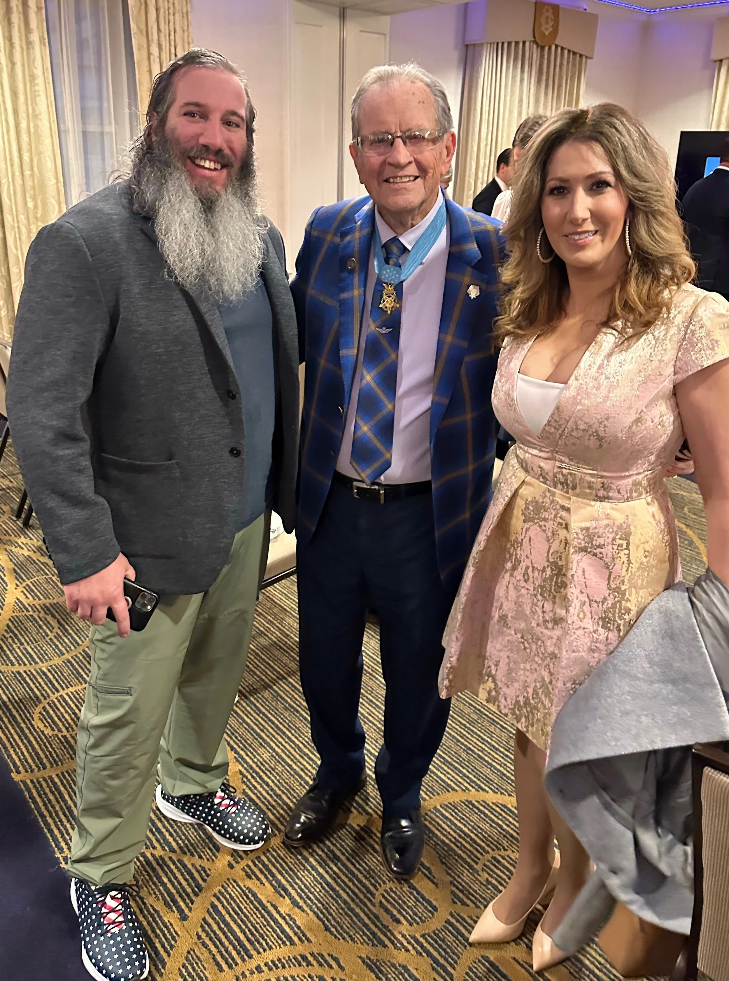 Three people posing for a photo at a formal event. The man on the left has a long beard, wearing a gray blazer, green pants, and polka dot sneakers. The man in the middle, wearing glasses, a blue plaid blazer with medals, and black shoes. The woman o