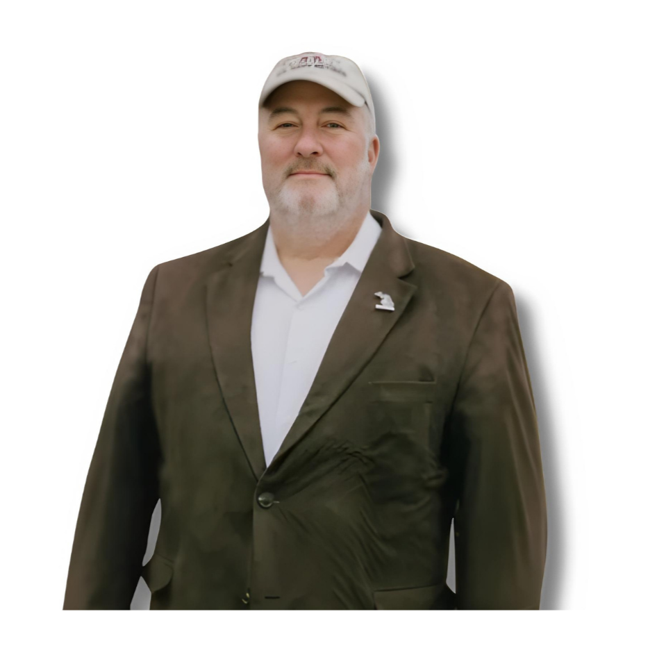 A middle-aged man wearing a white baseball cap, white dress shirt, and dark blazer with a lapel pin, standing against a plain white background.
