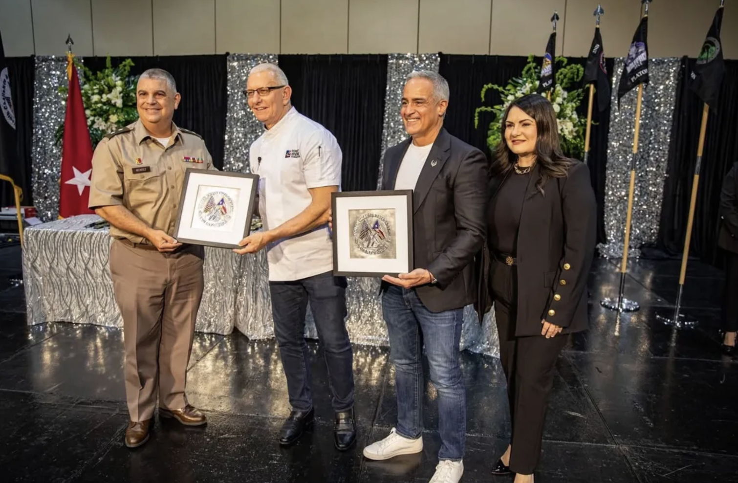 Four individuals, three men and one woman, standing on a stage during an award ceremony. The man on the left, in a military uniform, is holding a framed certificate. The second man from the left, in a white chef's coat, is also holding a framed certi