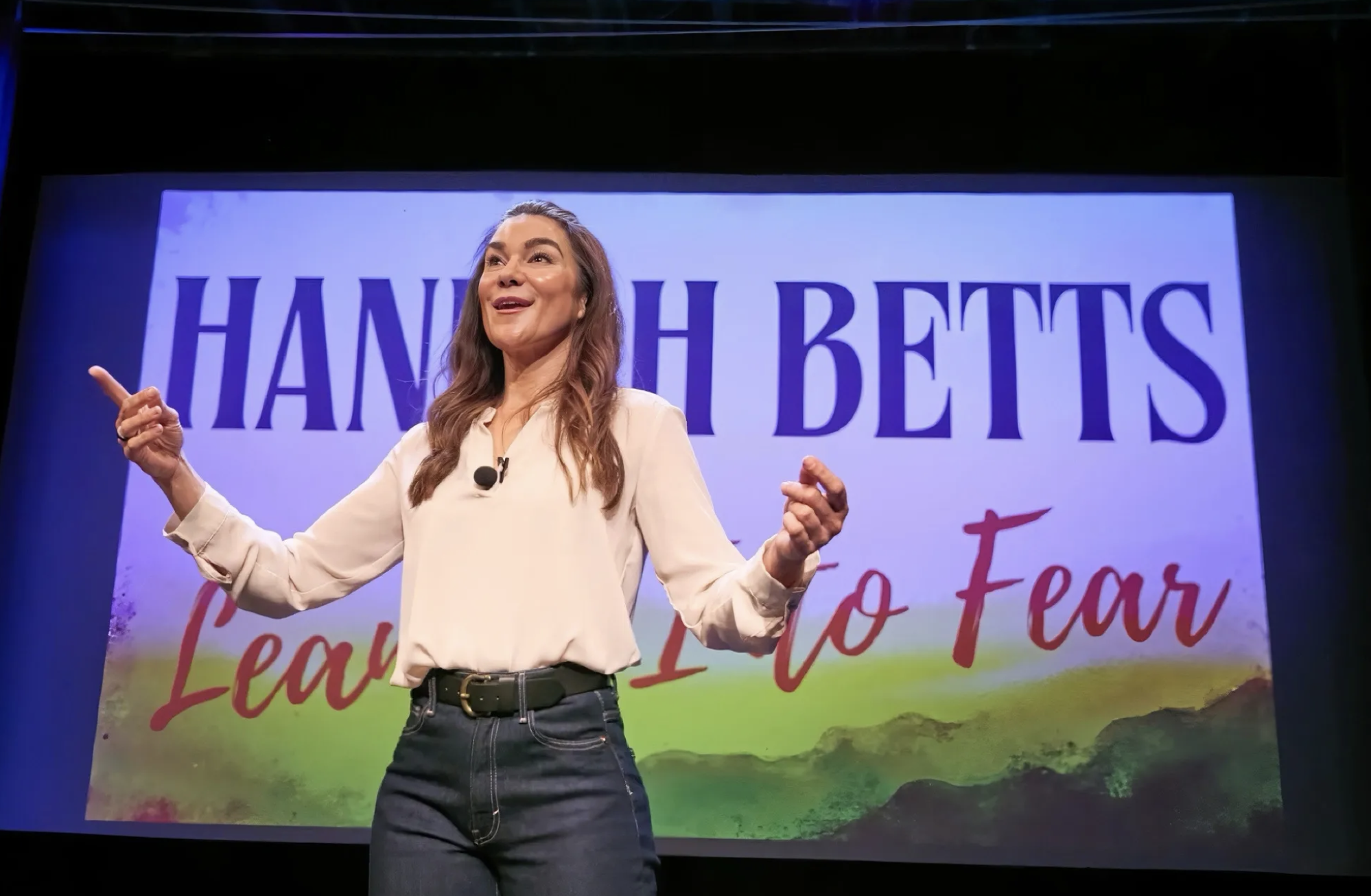 A woman standing in front of a colorful background with the words 'Hannah Bettis' and 'Fear to Fear', speaking or presenting with a microphone, gesturing with her hands.