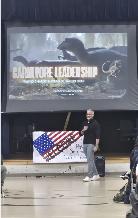 A man standing with a microphone in front of an audience, smiling, with a presentation slide behind him that says 'Carnivore Leadership' and features a dinosaur and a prehistoric animal, in a gymnasium or community center setting.