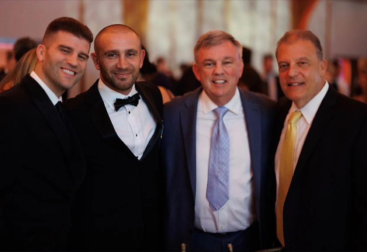 Four men wearing suits, standing close together and smiling at a formal event.