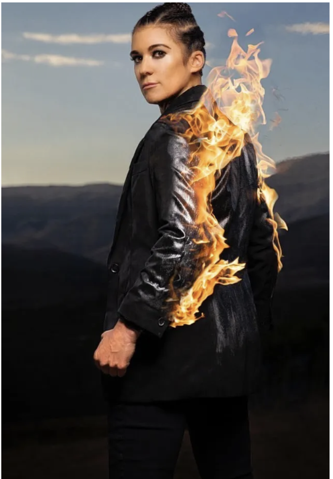 A woman standing outdoors with mountains in the background, wearing a black leather jacket, with fire seemingly emerging from her shoulder.