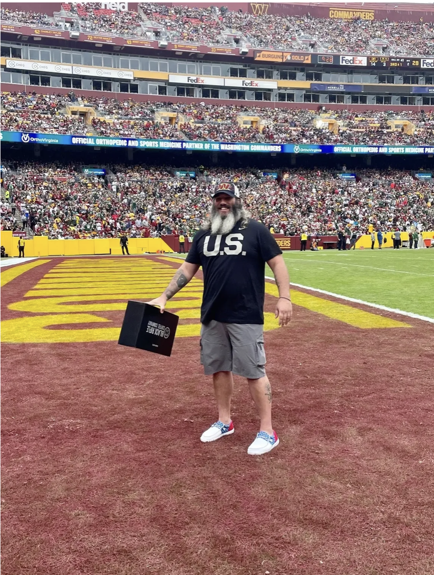 Man with long gray hair, beard, and tattoos on his legs, smiling on a football field, holding a box, wearing a black shirt with 'U.S.' printed on it and a 'W