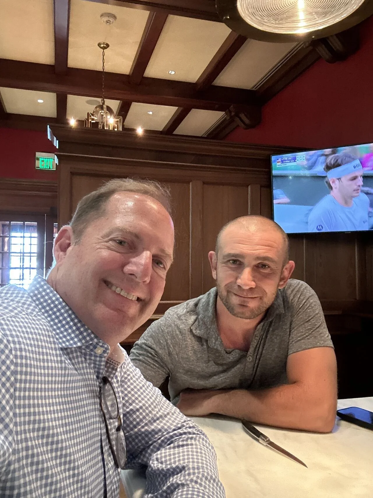 Two men sitting at a table in a restaurant. One is smiling and wearing a checkered shirt, the other is more serious and wearing a gray shirt. A TV is mounted on the wall behind them, showing a tennis match.