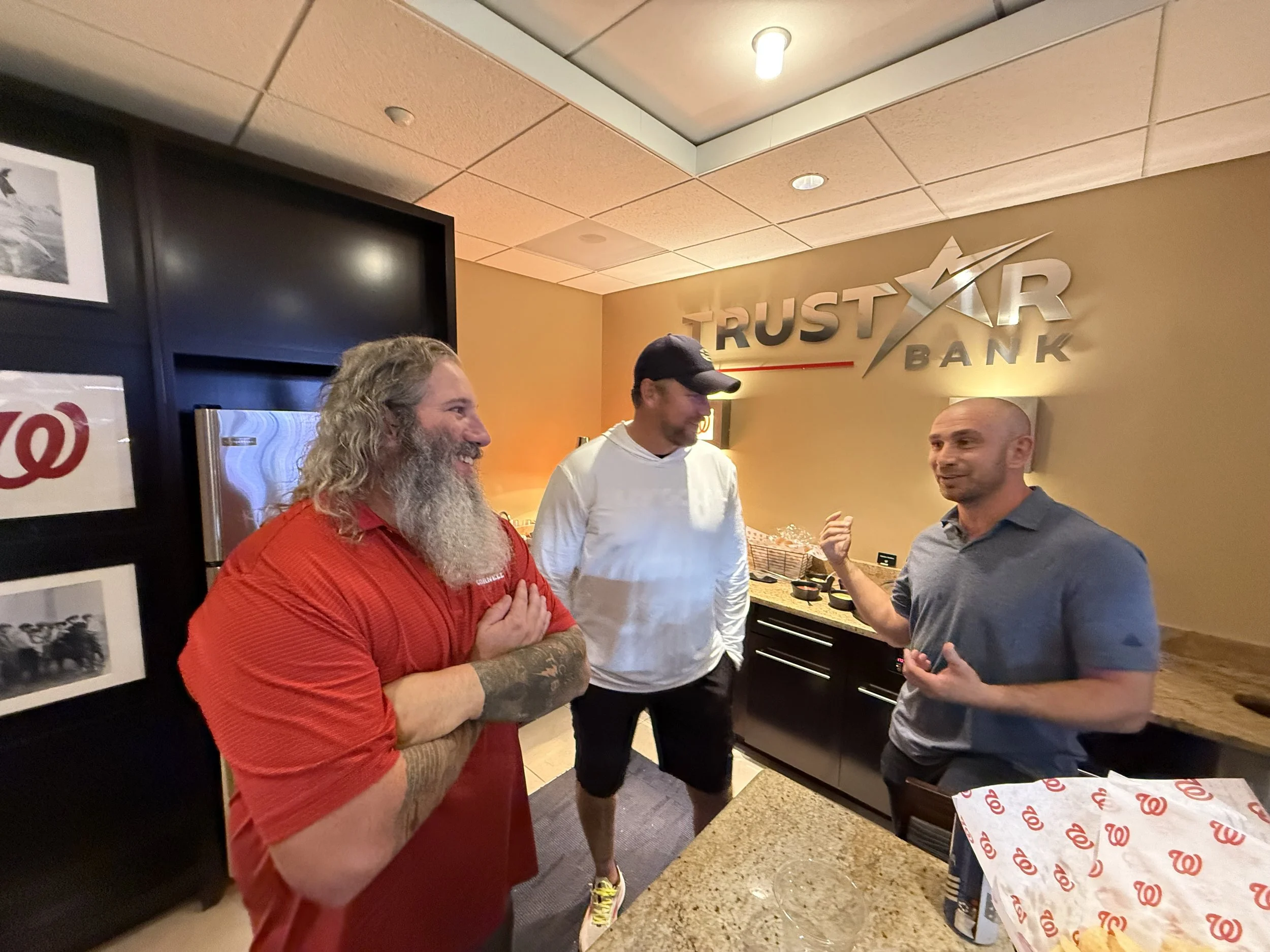 Three men standing and talking in a room with a sign on the wall that says "Trustar Bank." One man with long curly blonde hair and beard wearing a red shirt, another man with short dark hair and beard wearing a white shirt, and a third man with a sha