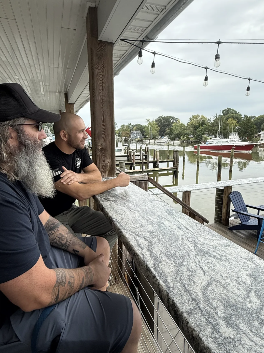 Two men sitting on a covered porch overlooking a boat dock and water, with parked boats and trees in the background, under a cloudy sky.