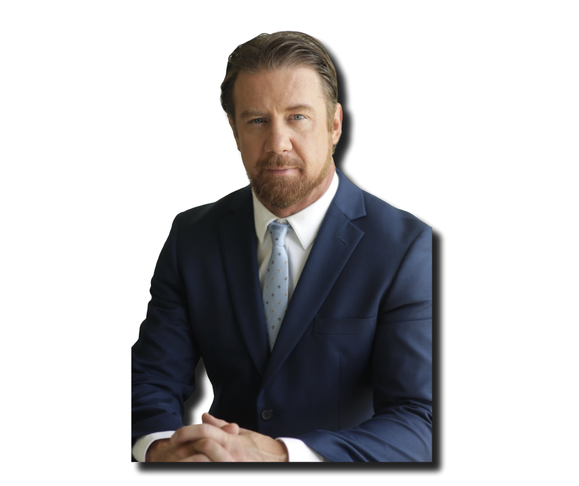 A man in a navy blue suit with a white shirt and light blue polka dot tie, sitting with hands clasped, against a black background.