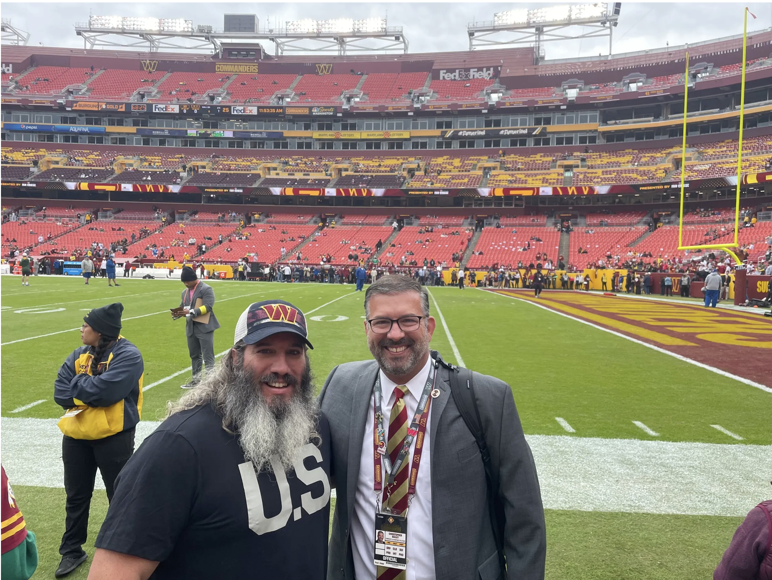 Two men smiling at a football stadium field, with one wearing a black U.S. football shirt and the other in a suit and tie. There are people and stadium seats in the background.