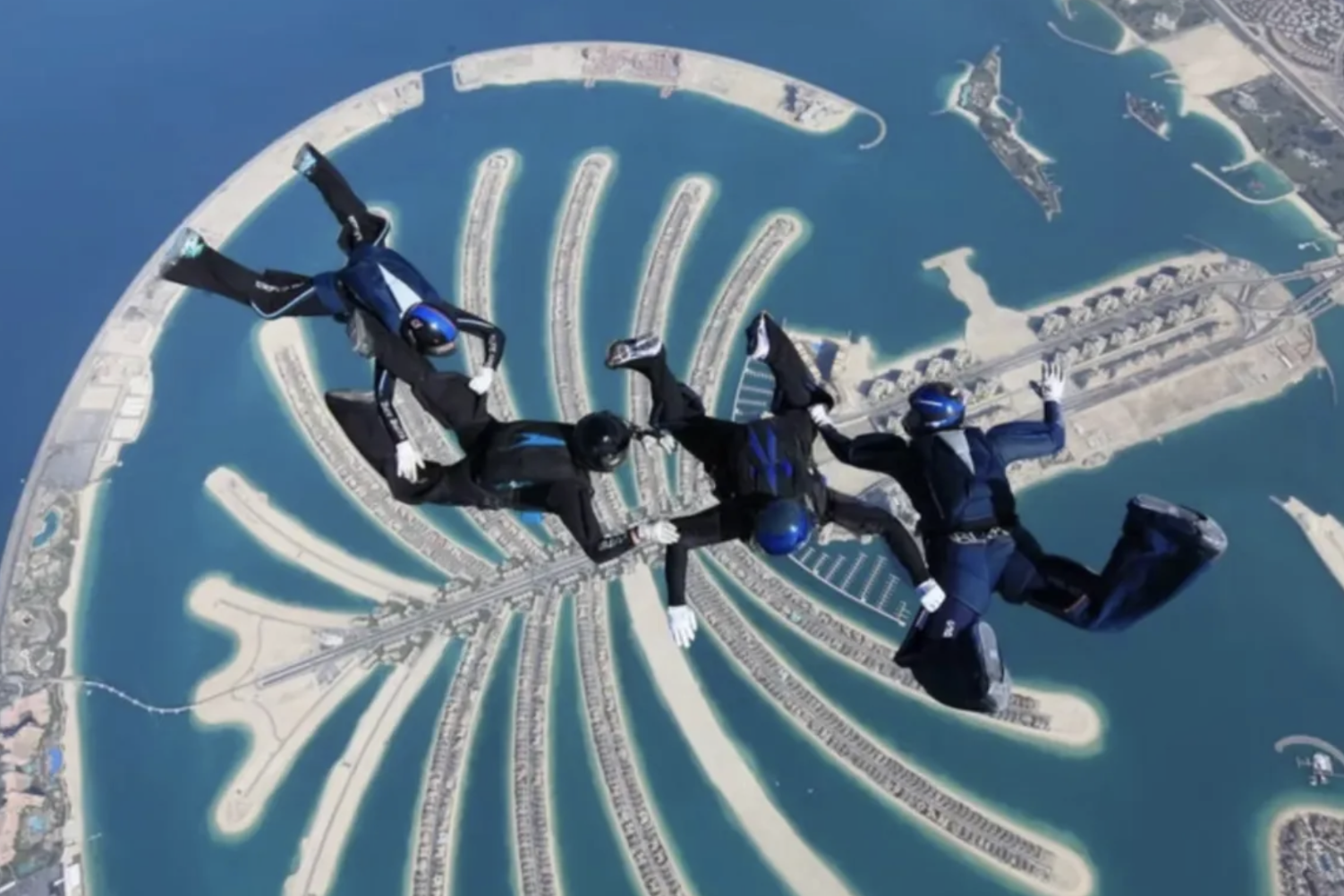 Four skydivers in black suits and blue helmets free-falling over the Palm Jumeirah, an artificial island in Dubai shaped like a palm tree surrounded by water with a marina and other islands visible.