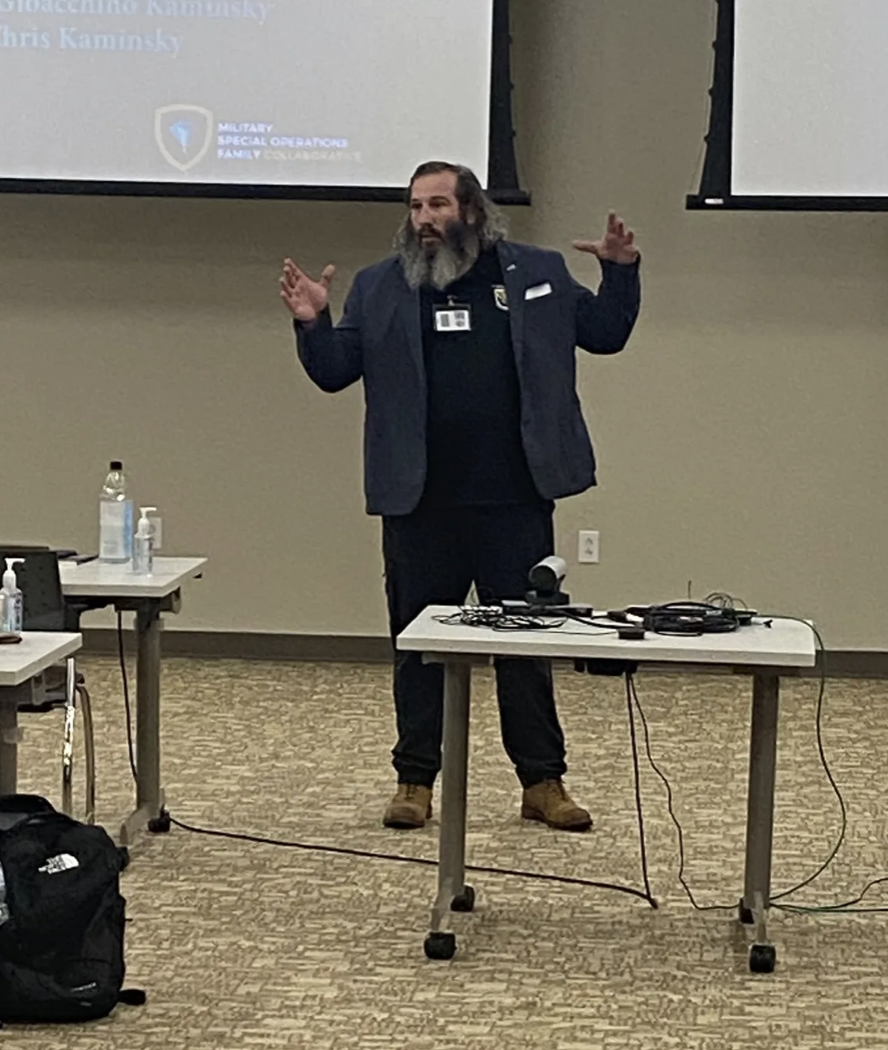 A man with a beard wearing a dark jacket and black shirt stands at a presentation in a conference room. He gestures with his hands while speaking. There are tables with bottles of hand sanitizer, electronic devices, and cables, with a large screen be