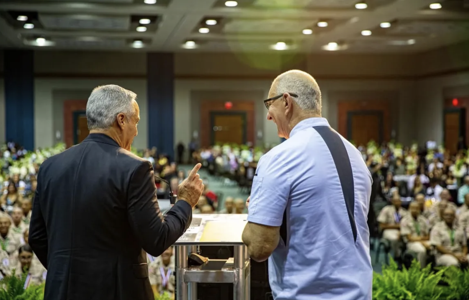Two men are speaking on a stage in front of a large seated audience in a conference hall. One man is wearing a dark suit, and the other is dressed in a light blue shirt with black glasses. The man in the suit is pointing a finger as they converse.