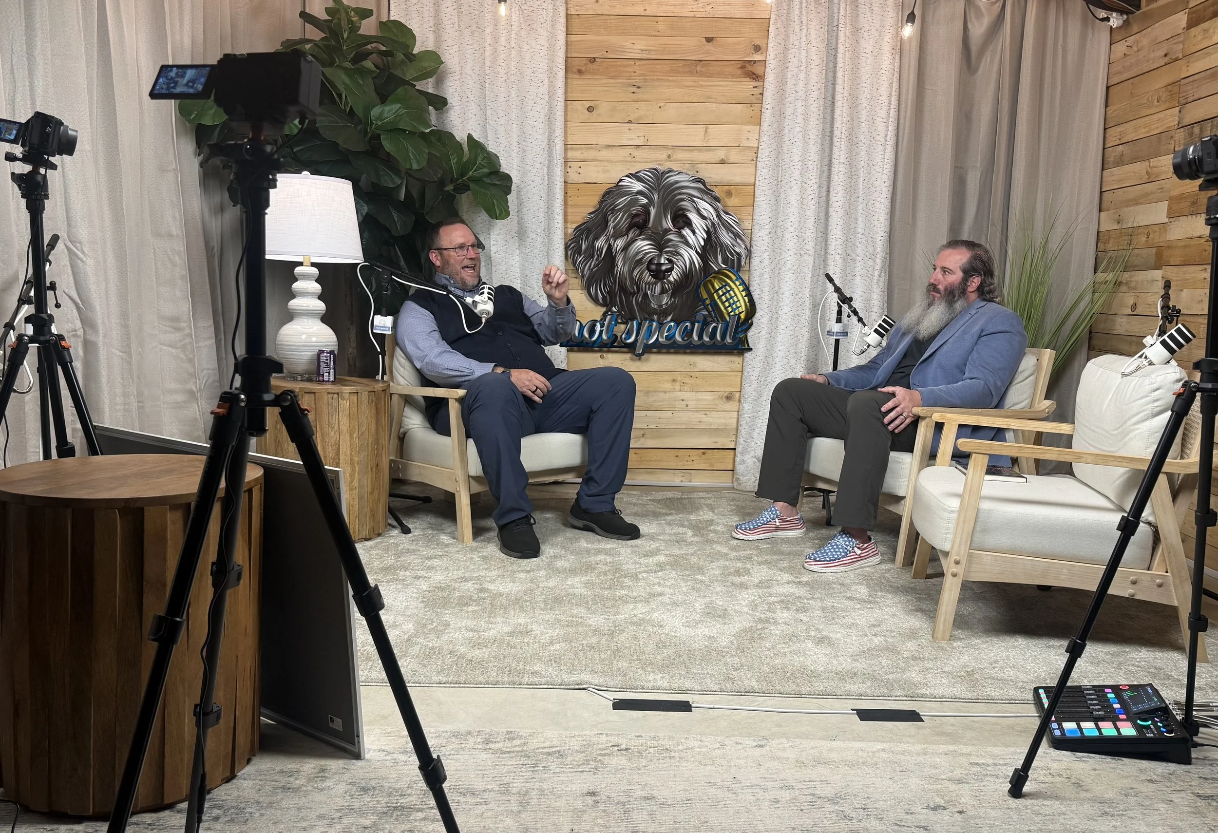 Two men are seated in a studio setting with a wood-paneled wall behind them. There are microphones on stands in front of them, and one man is speaking animatedly. The other man is listening. The studio is decorated with plants, a table lamp, and a si