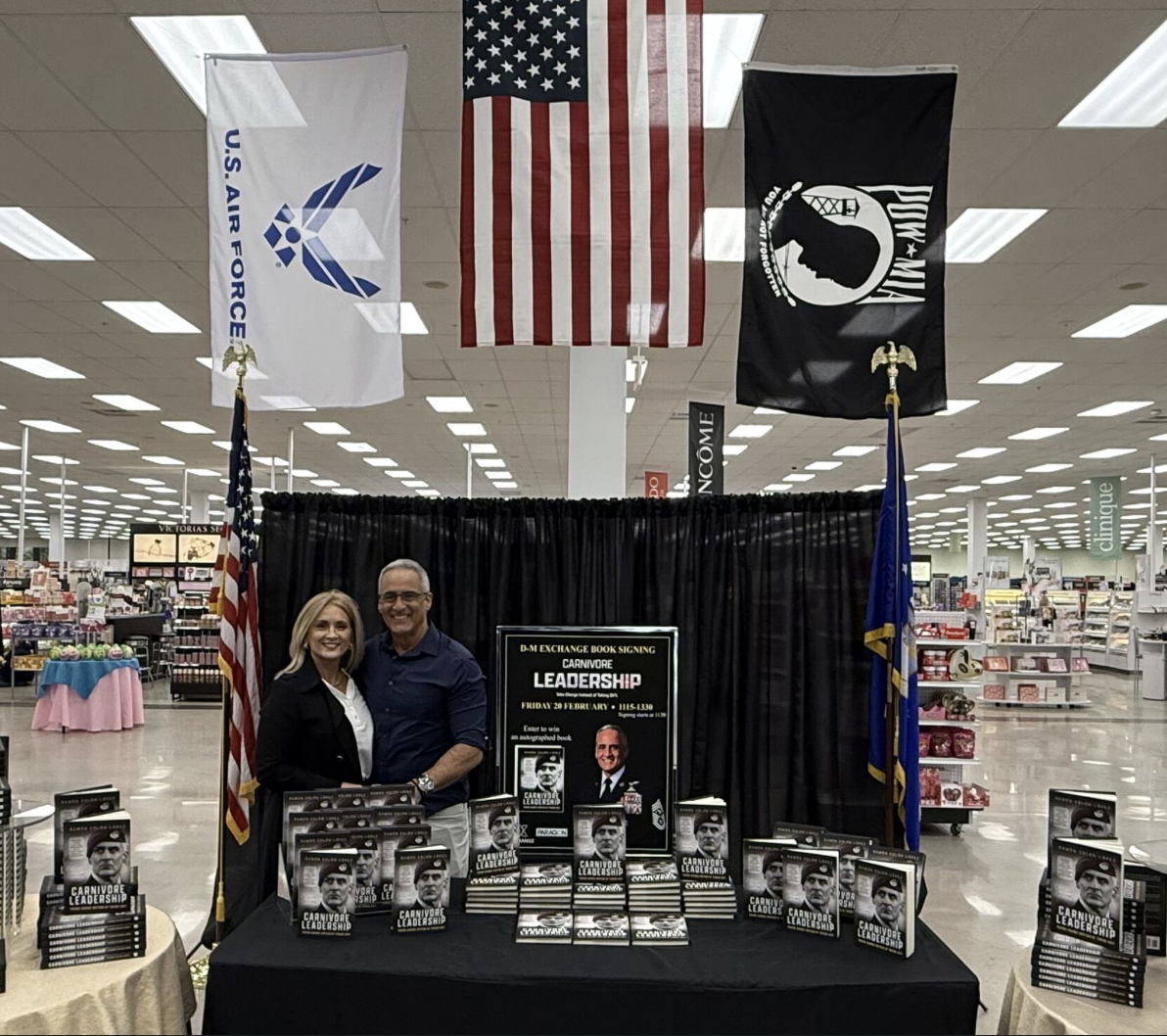A book signing event for 'Carnivore Leadership' taking place in a store. Two people are standing behind a table with multiple copies of the book, banners, and patriotic flags hanging above. The background shows store shelves.