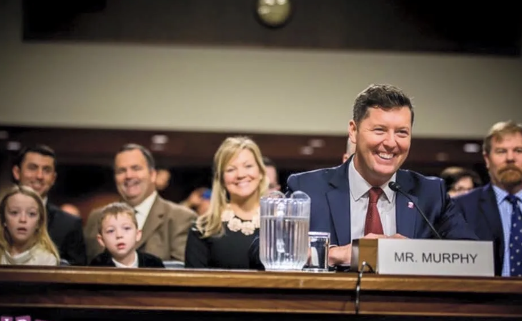 A group of people, including children and adults, sitting behind a official nameplate that reads "MR. MURPHY" at a formal hearing or meeting, smiling and listening attentively.
