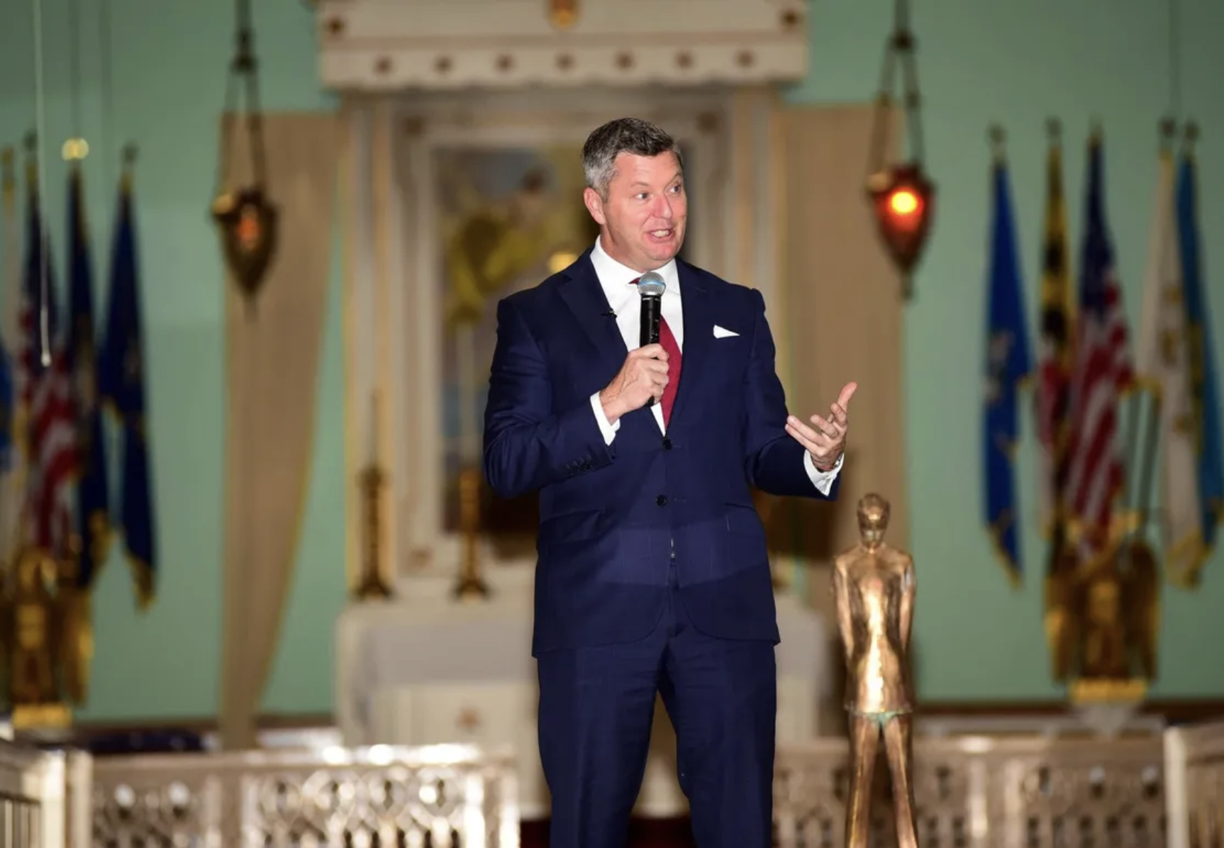 A man in a navy blue suit and red tie speaking into a microphone on stage, with flags in the background and a golden sculpture of a human figure nearby.