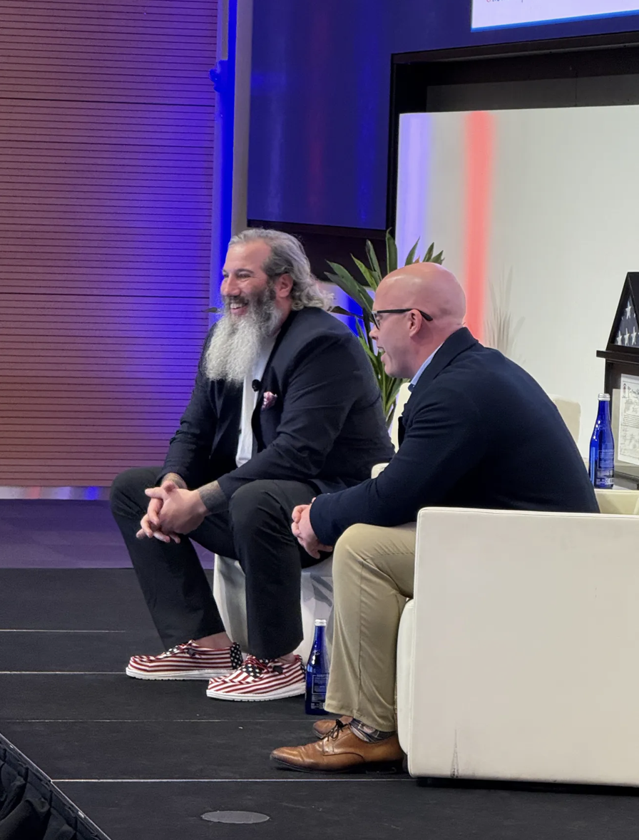 Two men seated on stage during a panel or interview, smiling and engaged in conversation. One man has a long gray beard and is wearing a dark blazer, white shirt, and red-and-white striped shoes. The other man is bald, wearing glasses, a dark blazer,