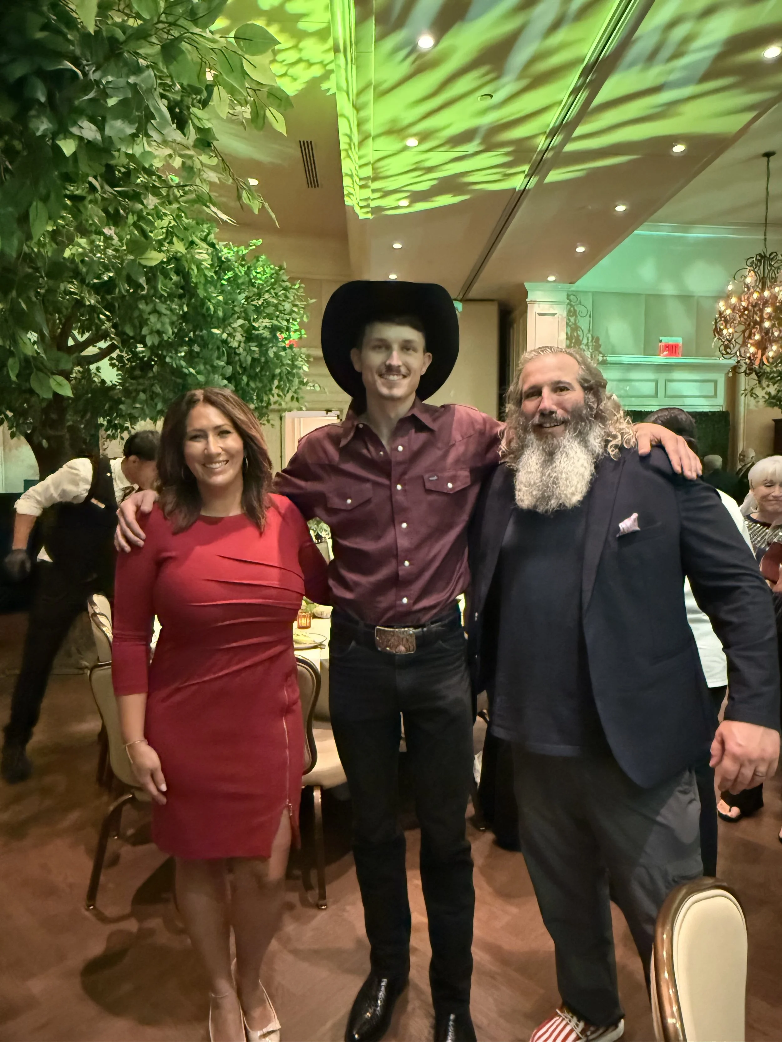 Three people standing together at a social event, indoors with green lighting, a large tree, and chandelier decorations. The group includes a woman in a red dress, a young man with a black hat and burgundy shirt, and a man with a gray beard in a dark