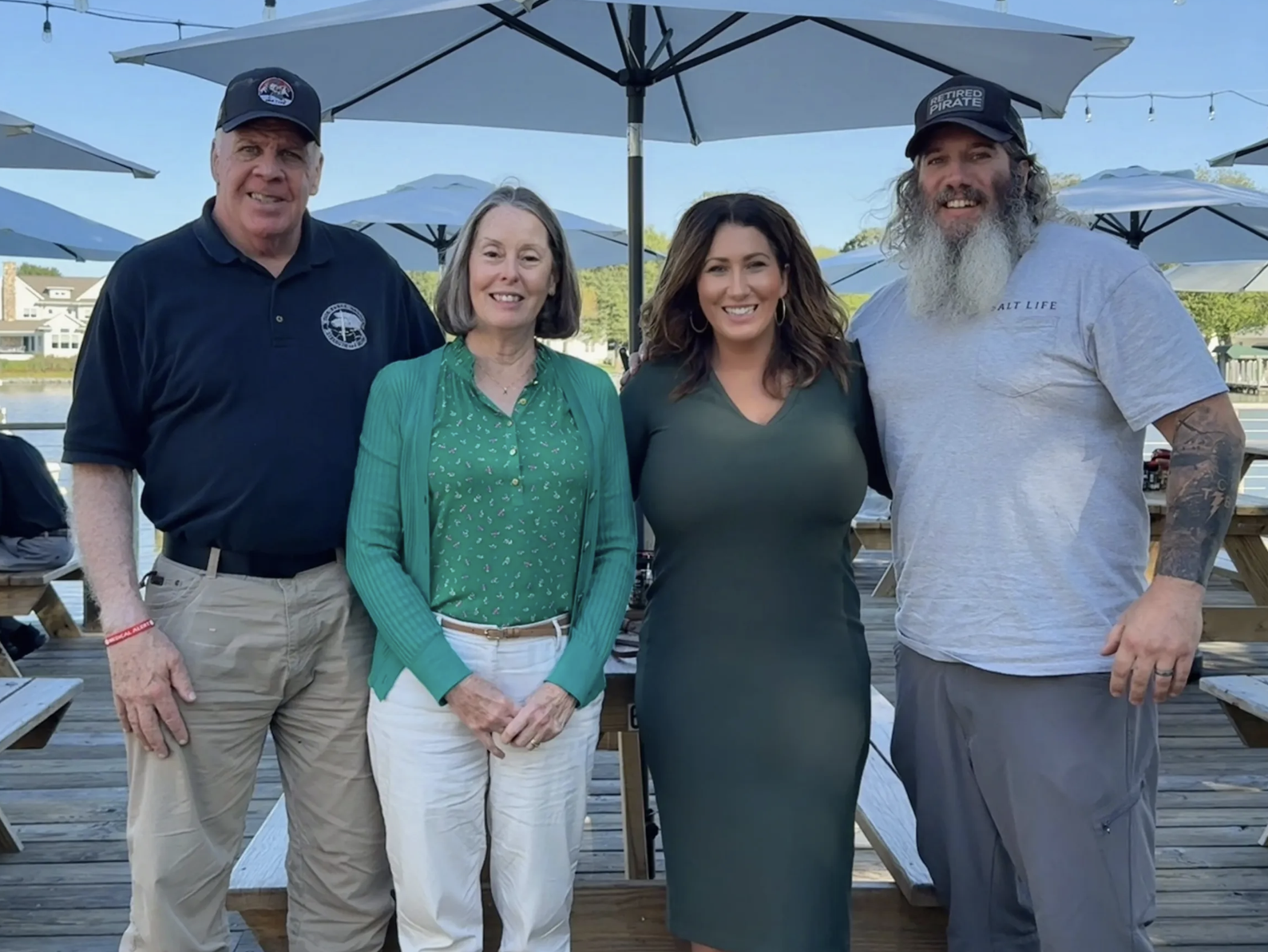 Four people standing on a deck outdoors, with umbrellas and a body of water in the background, smiling at the camera.