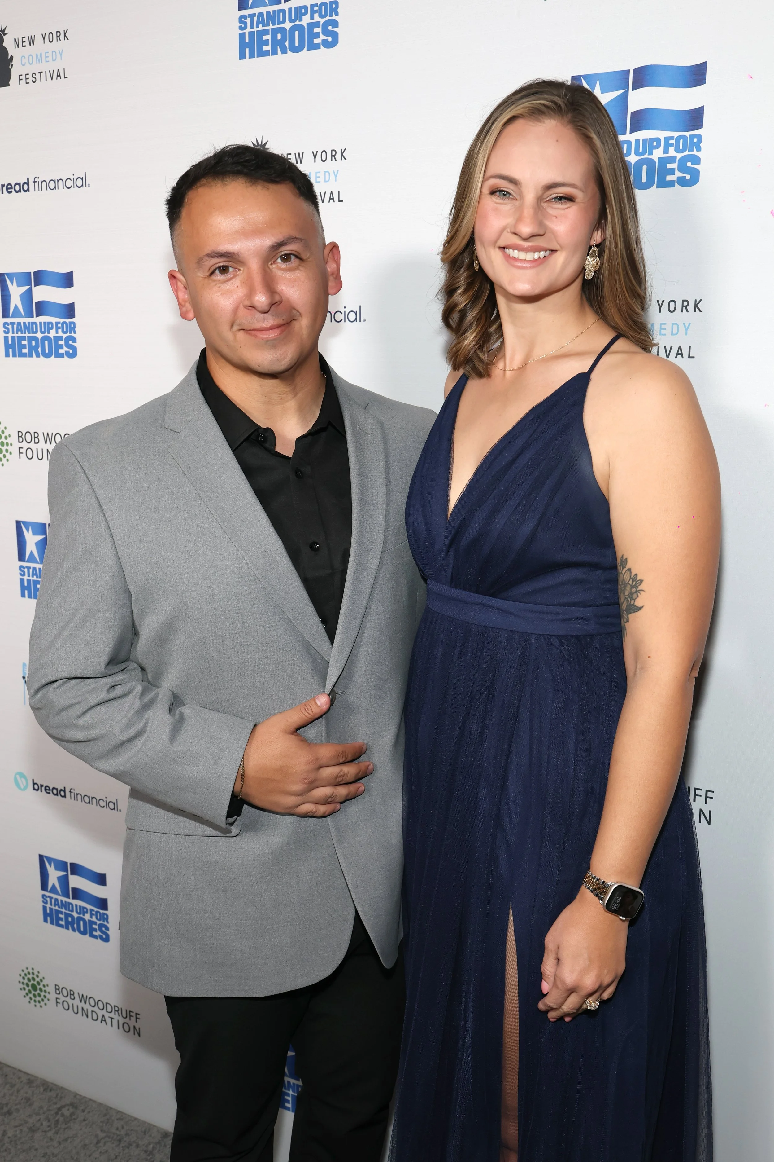 A man and a woman standing together at an event, with a backdrop featuring logos and text for the 'Stand Up for Heroes' event and the New York Comedy Festival. The man is wearing a light gray suit with a black shirt, and the woman is wearing a dark blue sleeveless dress with earrings and a watch. Both are smiling at the camera.