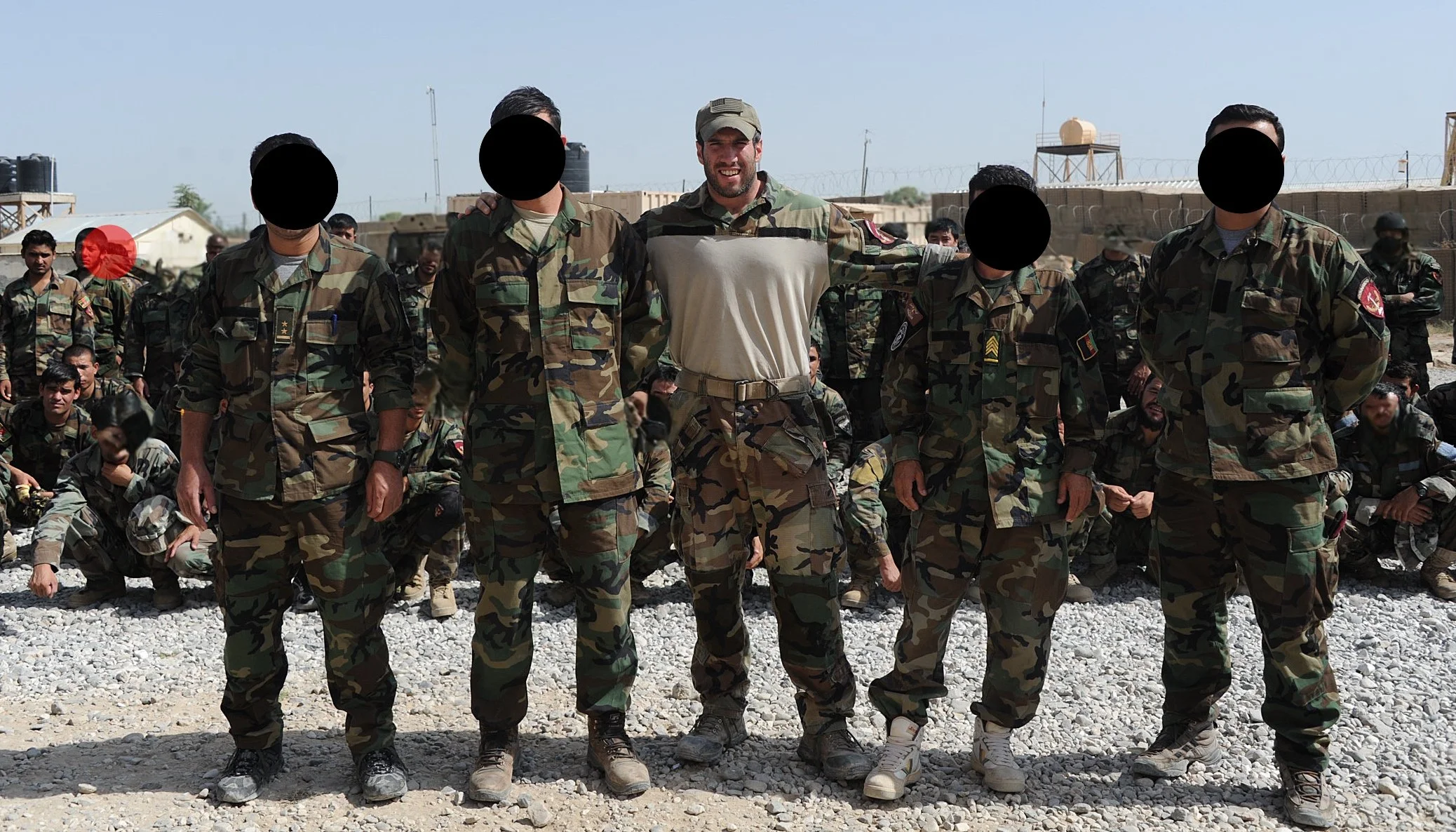 Group of soldiers in camouflage uniforms standing outdoors on rocky ground, with some seated behind, and a military base with barbed wire and water tanks in the background.