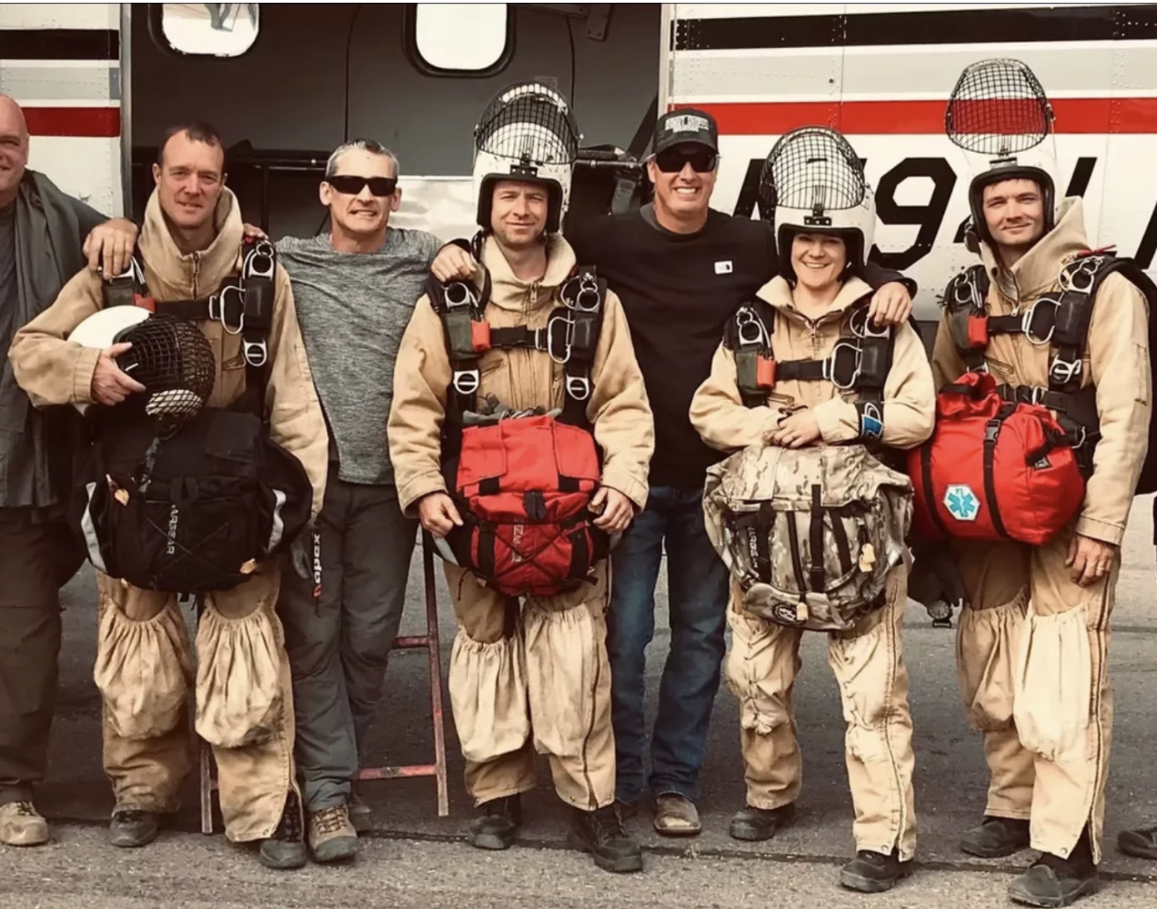 Group of five firefighters in tan uniforms with helmets and gear, standing with two civilian men in front of a fire truck.
