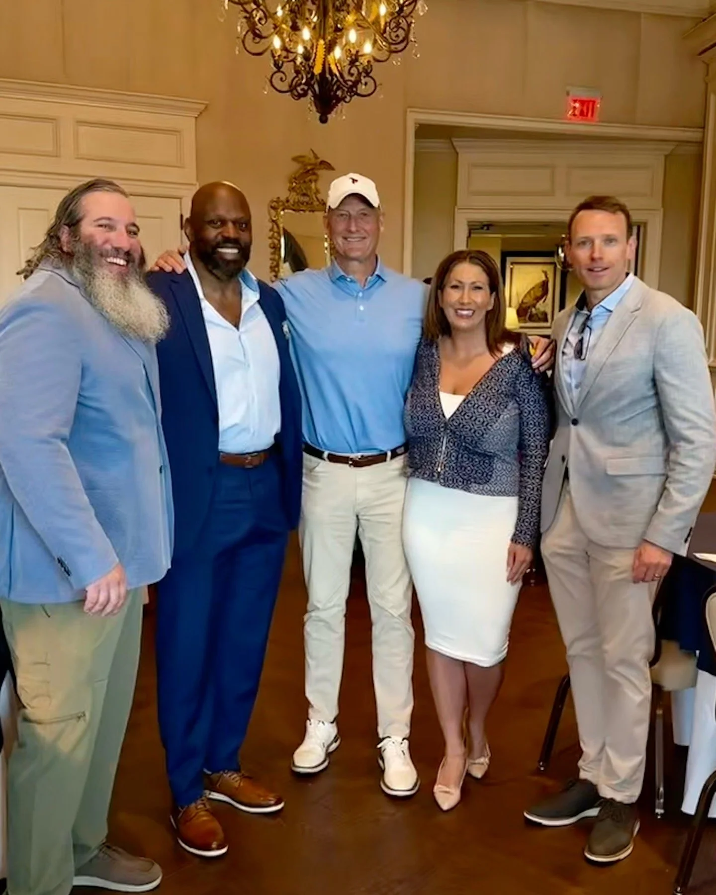 A group of six people standing together in a warmly lit room, smiling for a photo. The group includes five men and one woman, dressed in smart casual attire, with one man wearing a white cap. The background features elegant decor, including a chandel
