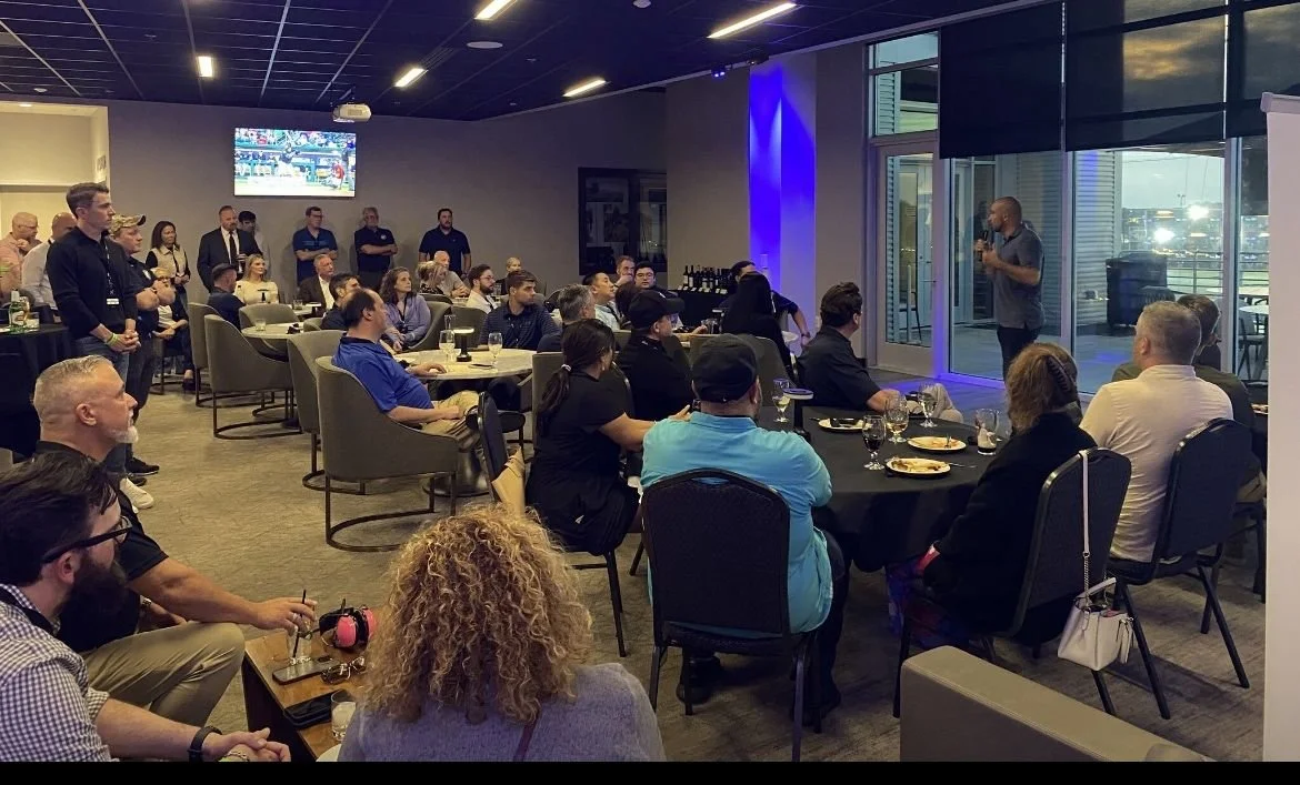 A man speaks to an audience seated at round tables in a conference room with large windows, some standing against the wall and a TV screen on the wall.