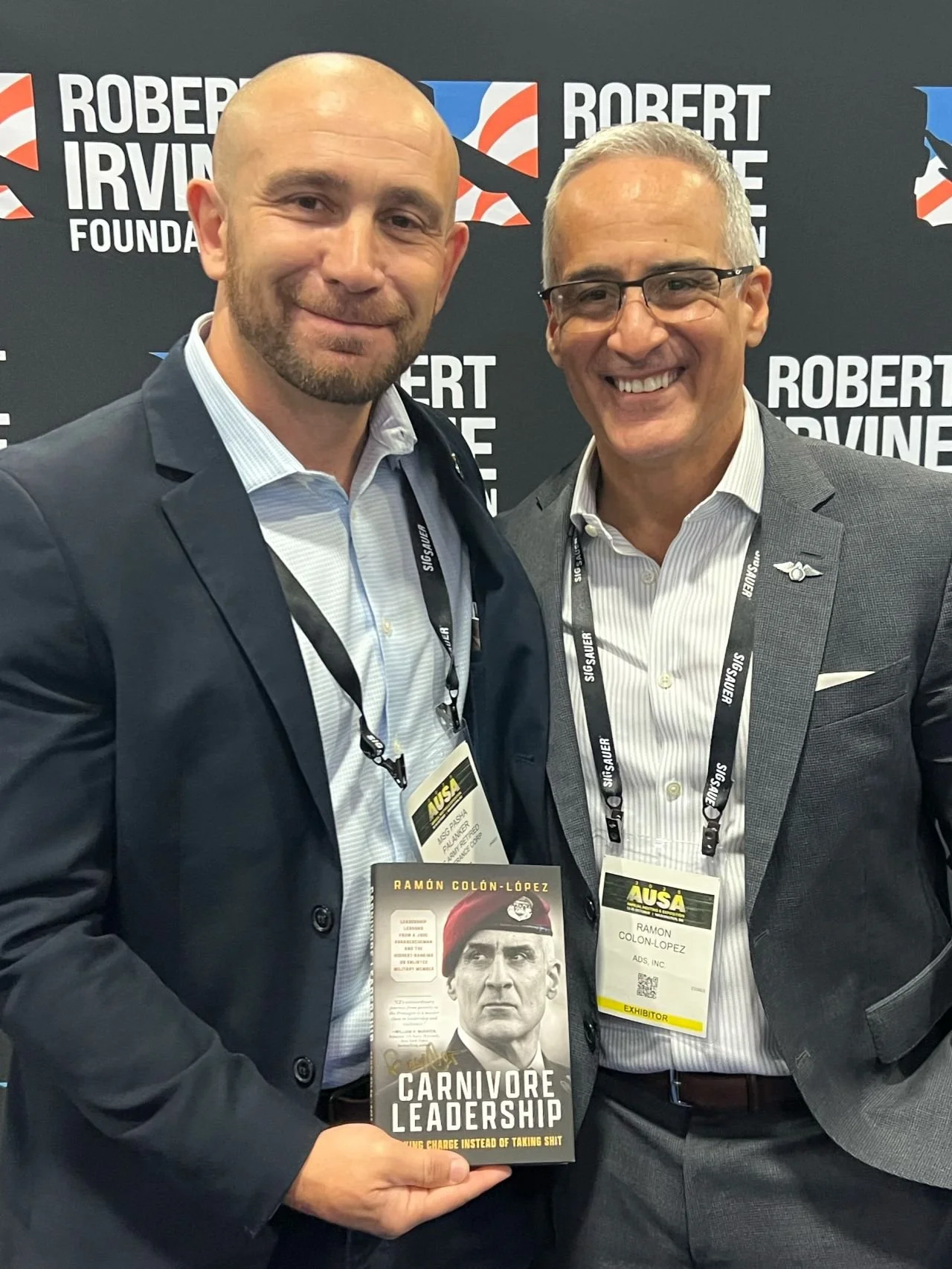 Two men in suits at a conference, one holding a book titled 'Carnivore Leadership' featuring a serious man in a red cap on the cover. Behind them is a backdrop with the name 'Robert Irvine Foundation'. Both men are wearing conference badges and smili