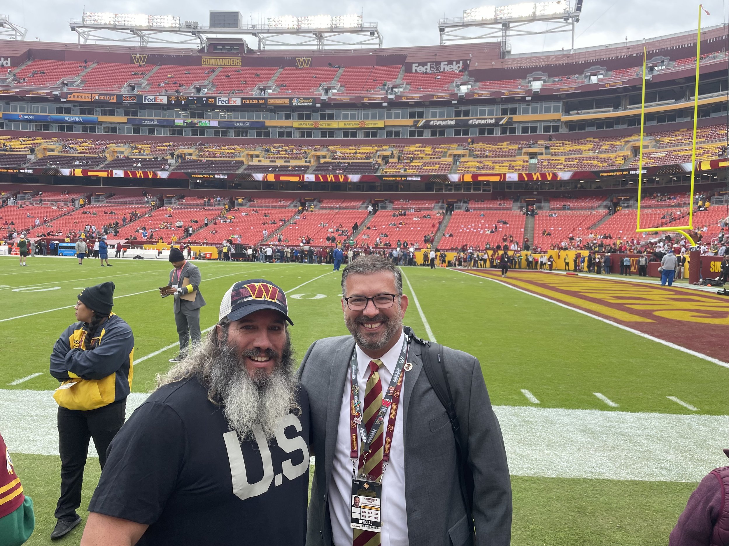 Two men smiling on a football field with stadium seating in the background. One is wearing a hat with a logo, and the other is dressed in a suit with a tie. The stadium seats are mostly empty.