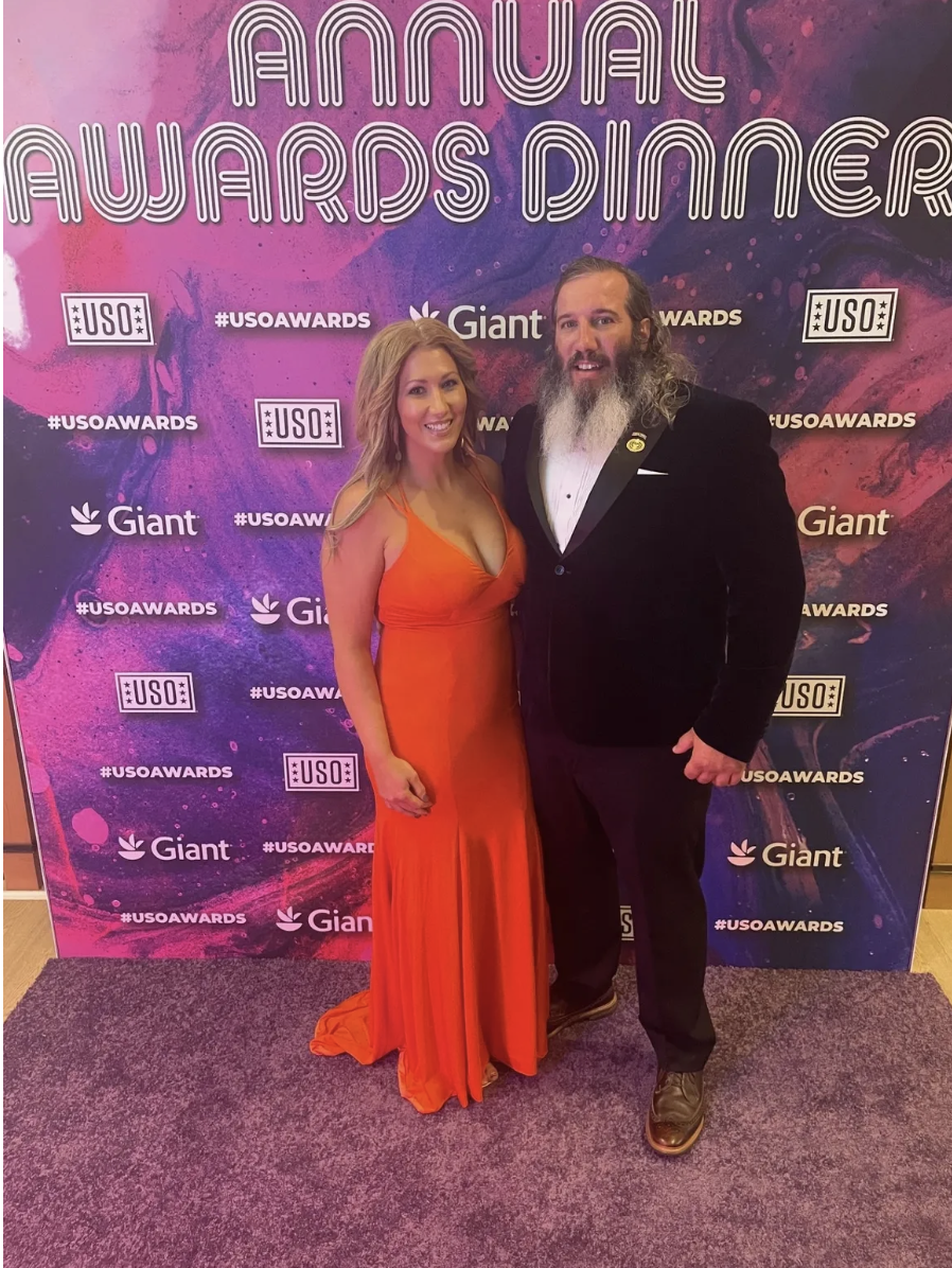 A woman in an orange gown and a man in a black tuxedo posing together at the USA Awards Dinner, with a vibrant pink and purple backdrop featuring the event's text and logos.