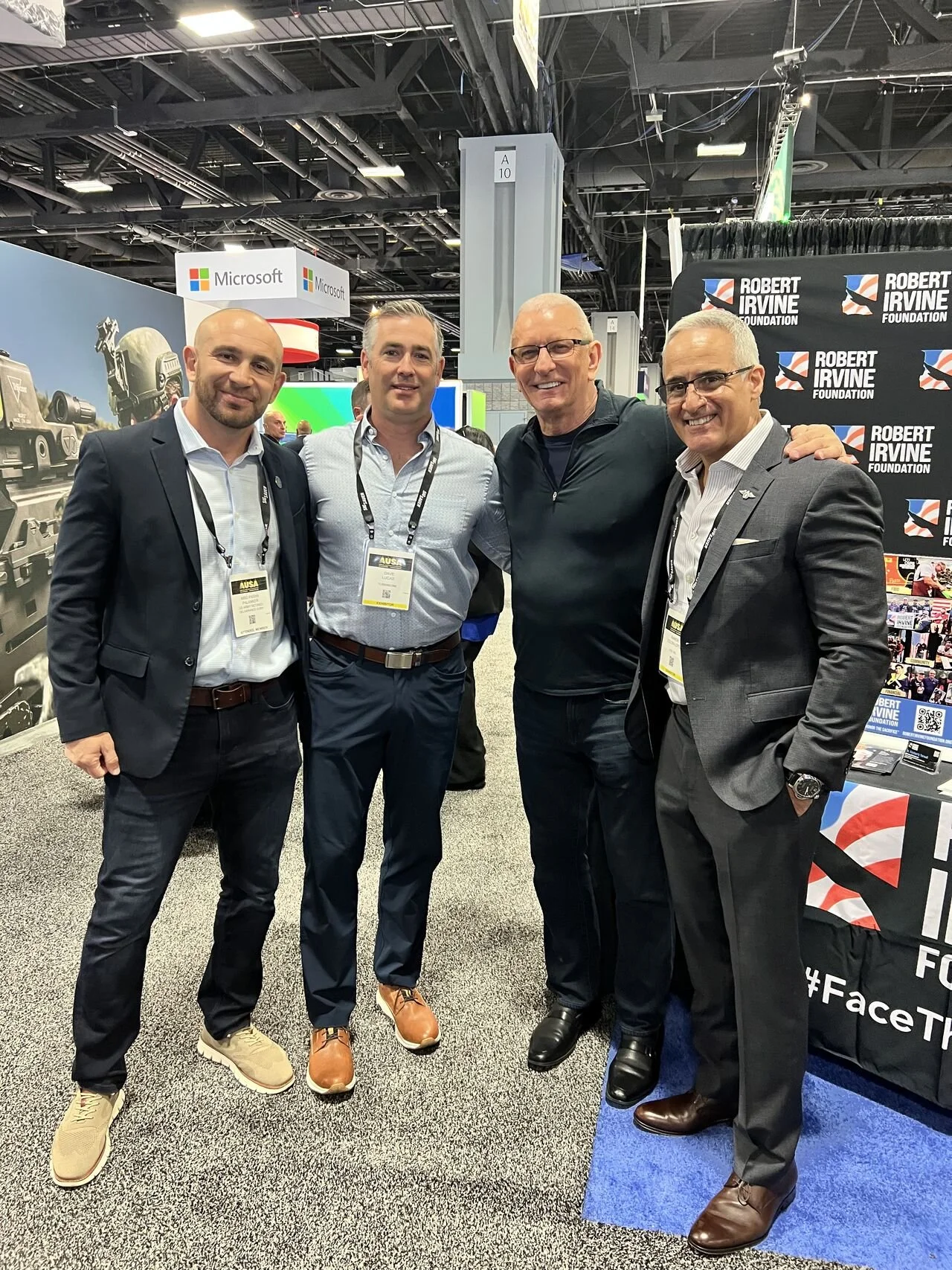 Four men standing together at a conference or event, smiling for the camera. They are dressed in business attire, with name badges hanging around their necks. The background shows booths with banners for Microsoft and the Robert Irvine Foundation.